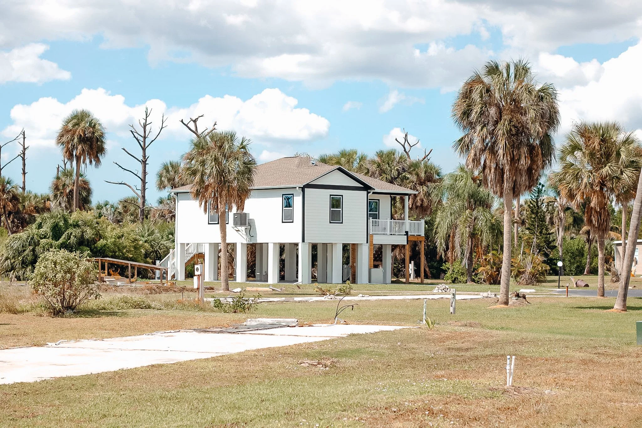 A white elevated house with black trim surrounded by palm trees, under a partly cloudy sky.