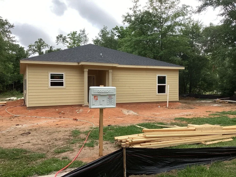 A house under construction with beige siding, two windows, and a dark gray roof, surrounded by dirt and construction materials, with trees in the background.