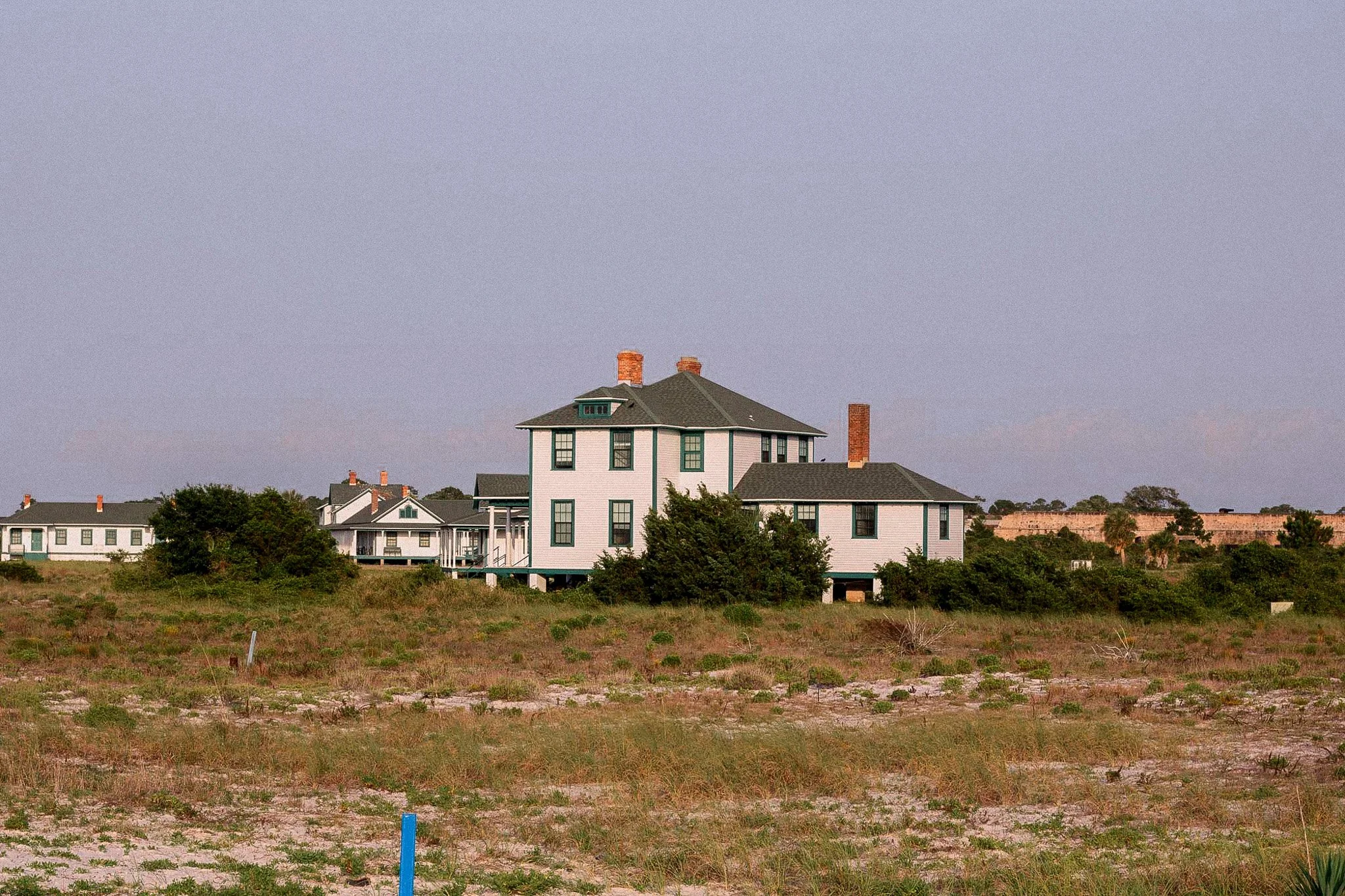 Large white house on a grassy, sandy plot of land with other similar houses in the background, under a clear sky.