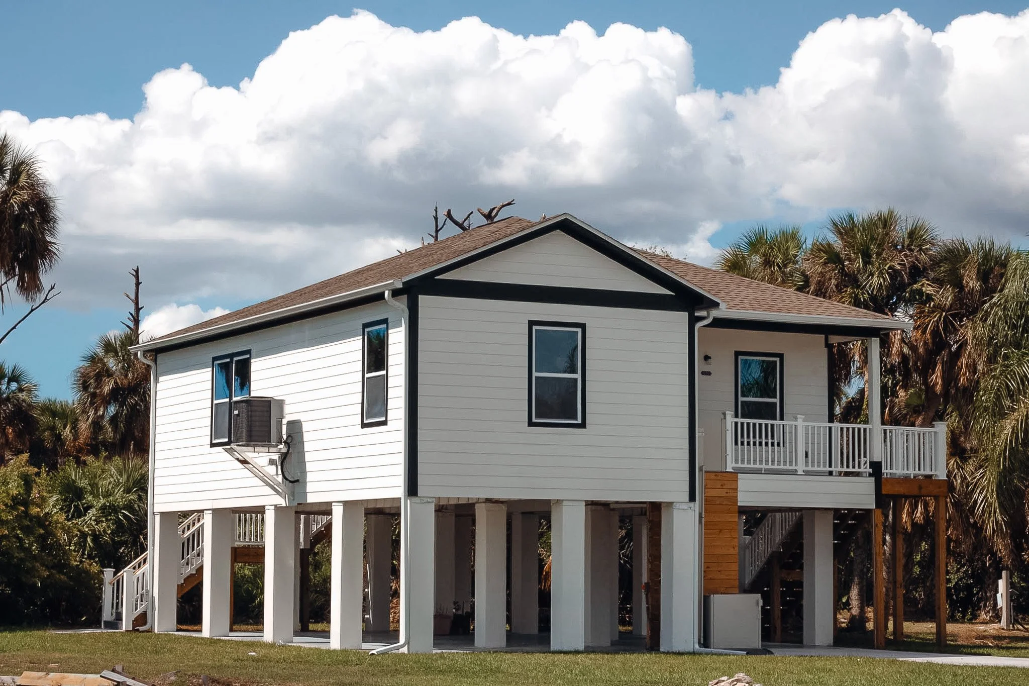 A two-story house elevated on stilts with white siding, black trim, a small balcony, and surrounded by palm trees under a partly cloudy sky.