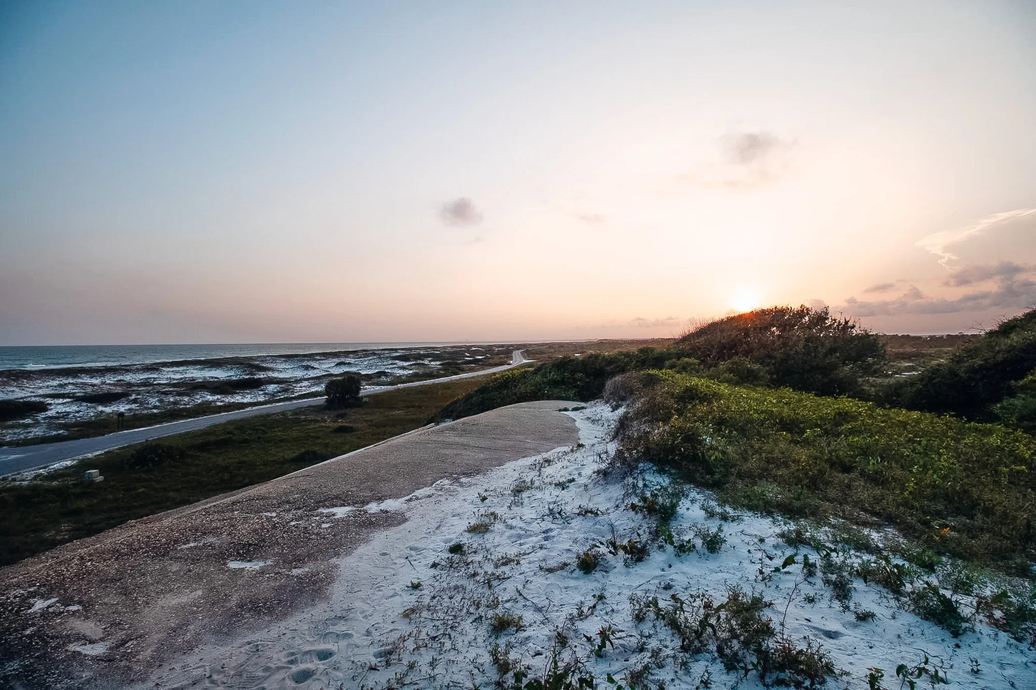 A sandy coastal landscape with a dirt path, grassy vegetation, and a sunset sky with clouds and the sun near the horizon.
