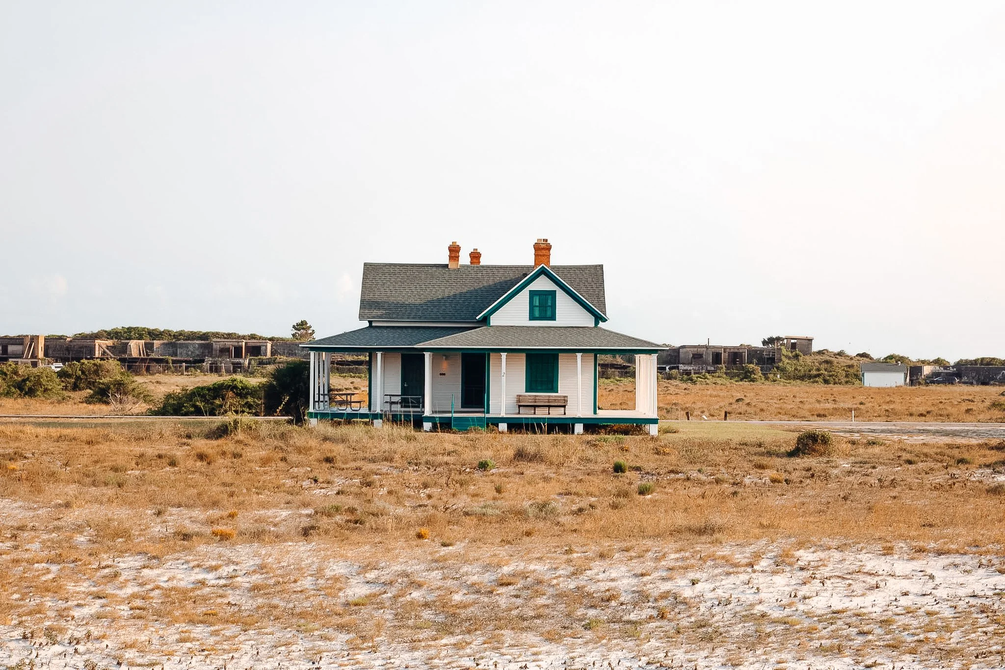A house with a gray roof, white walls, blue trim, by a dry, sandy landscape under a cloudy sky.