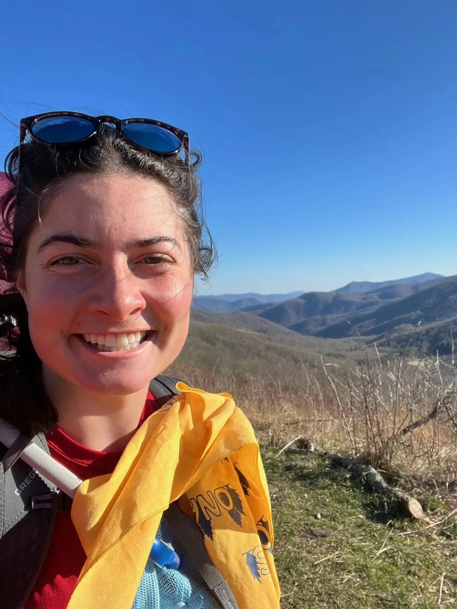 A woman smiling outdoors during a sunny day with mountains and dry grass in the background.