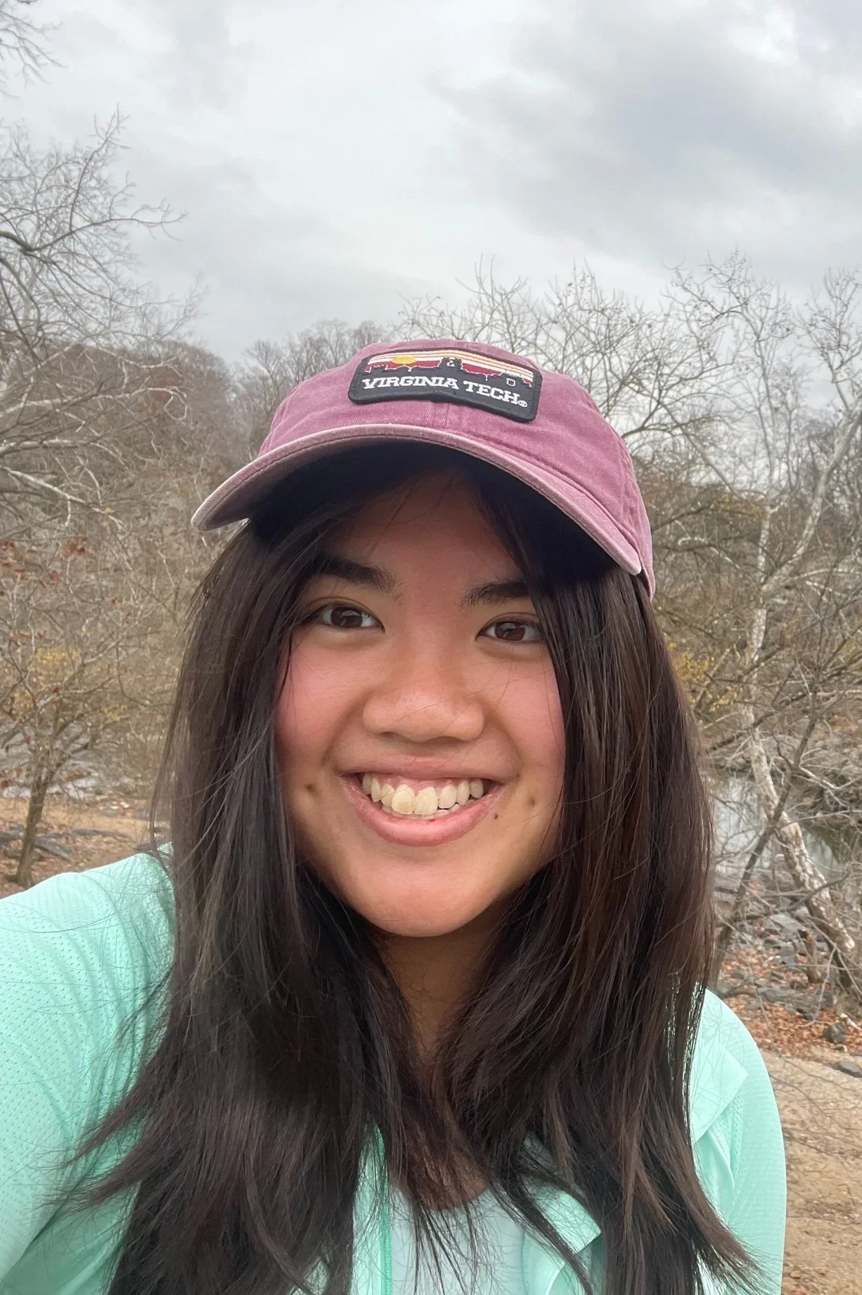 A young woman outdoors with a background of leafless trees and overcast sky, wearing a pink Virginia Tech cap and a light teal jacket, smiling at the camera.