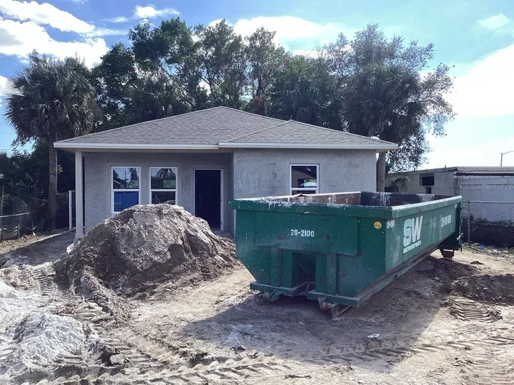 A house under construction with a large pile of dirt and an empty green dumpster in front.