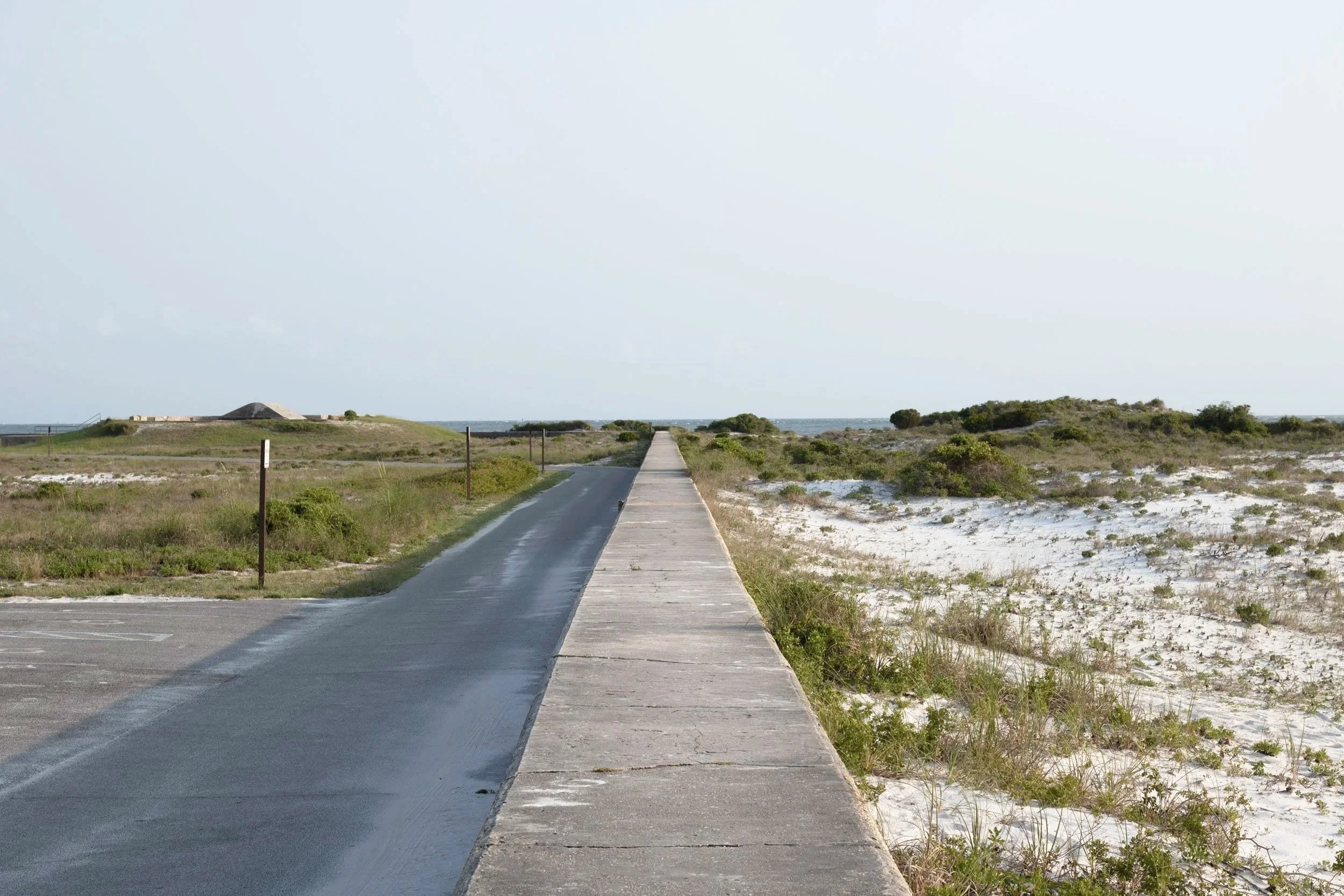 A paved road next to a concrete walkway running into a grassy, sandy landscape with sparse bushes and small trees, leading towards the horizon under a clear sky.