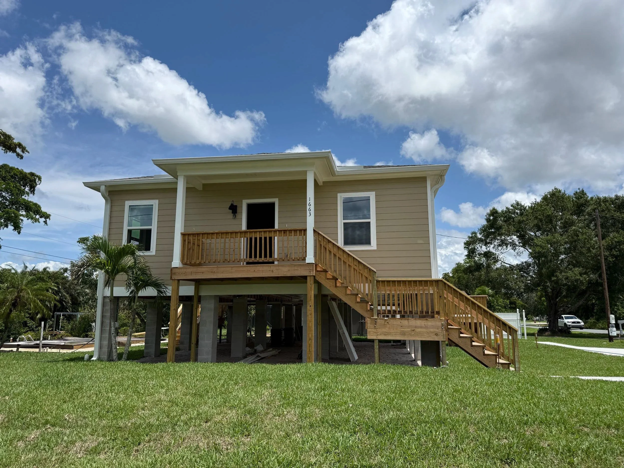 A two-story house with a beige exterior, white trim, and an unfinished wooden balcony and staircase. The house is elevated with open space underneath supported by concrete pillars. There is a small palm tree in the front yard, a green lawn, and some trees in the background. The sky is partly cloudy.