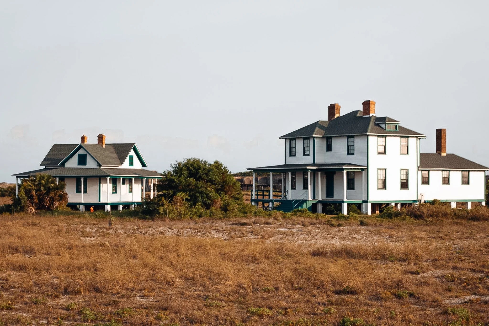 Two large white houses with black roofs and multiple chimneys sit on a grassy, dry field under a cloudy sky.