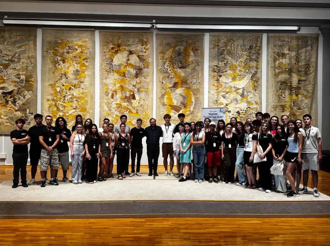 Group of young people and adults standing on a stage in front of large abstract woven textile artworks. The setting appears to be an indoor gallery or conference space, with a sign in the background that reads 'Tŭdarmai Bumi'.
