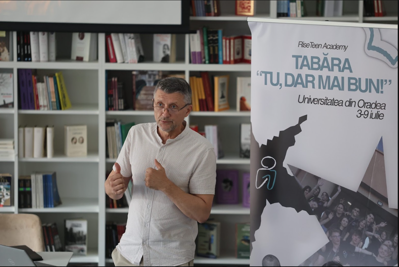 A man with glasses and gray hair gestures with his thumbs up while standing in front of a bookshelf and a banner promoting a university event in Oradea, Romania, with the date July 3-9.