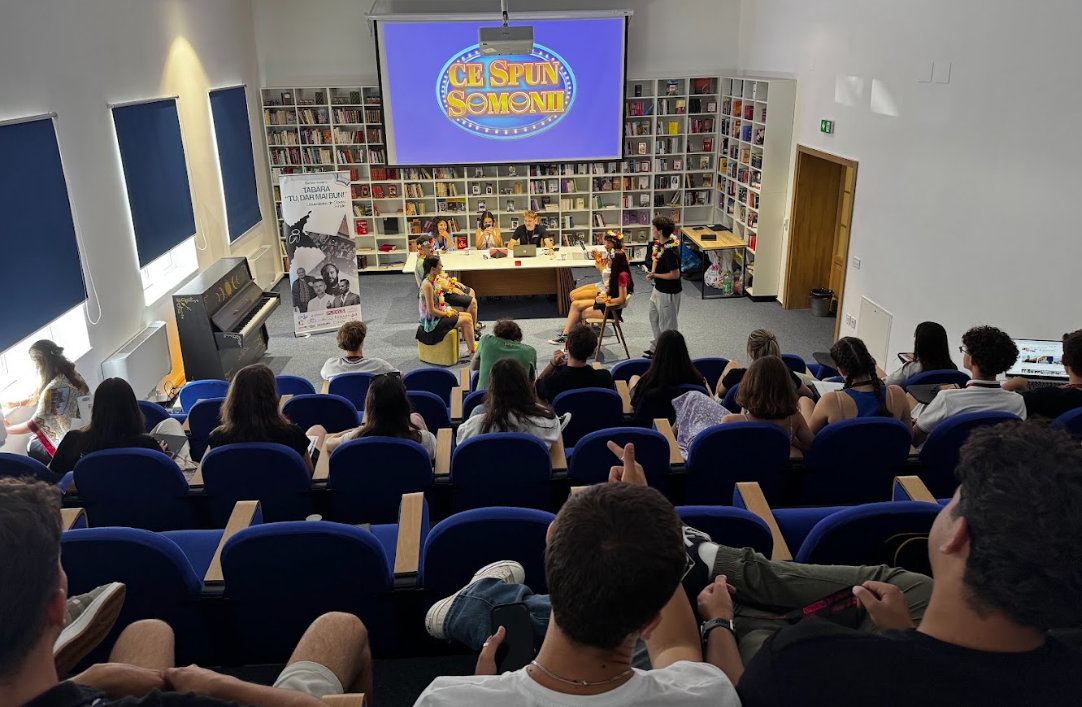 Children performing a theatrical play on stage in front of an audience in a library or bookstore, with bookshelves and a large screen displaying a colorful logo.