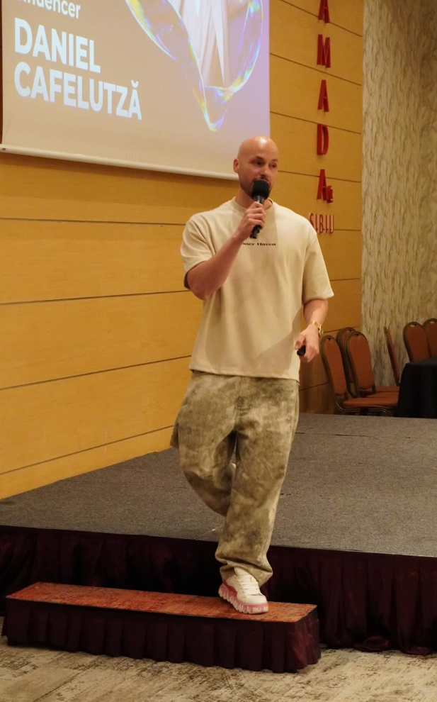 A man holding a microphone speaking on a stage with a large screen behind him displaying his name, Daniel Cafelutza, and a heart-shaped graphic. He is dressed in a beige oversized t-shirt, camouflage pants, and white sneakers, standing with one foot on an elevated platform.
