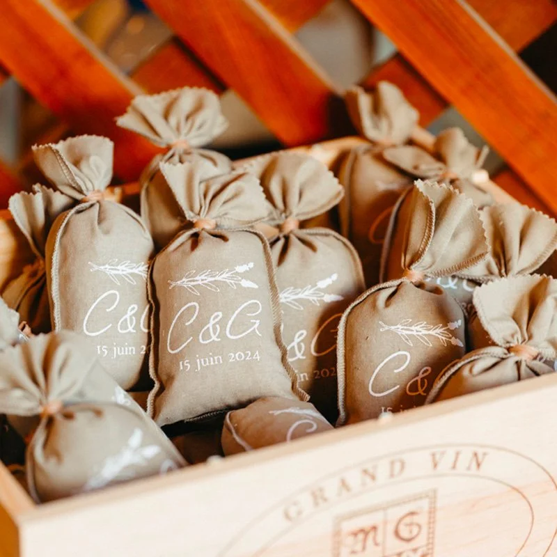 Petites pochettes en tissu beige avec inscription et date, dans une caisse en bois, avec un fond en bois.