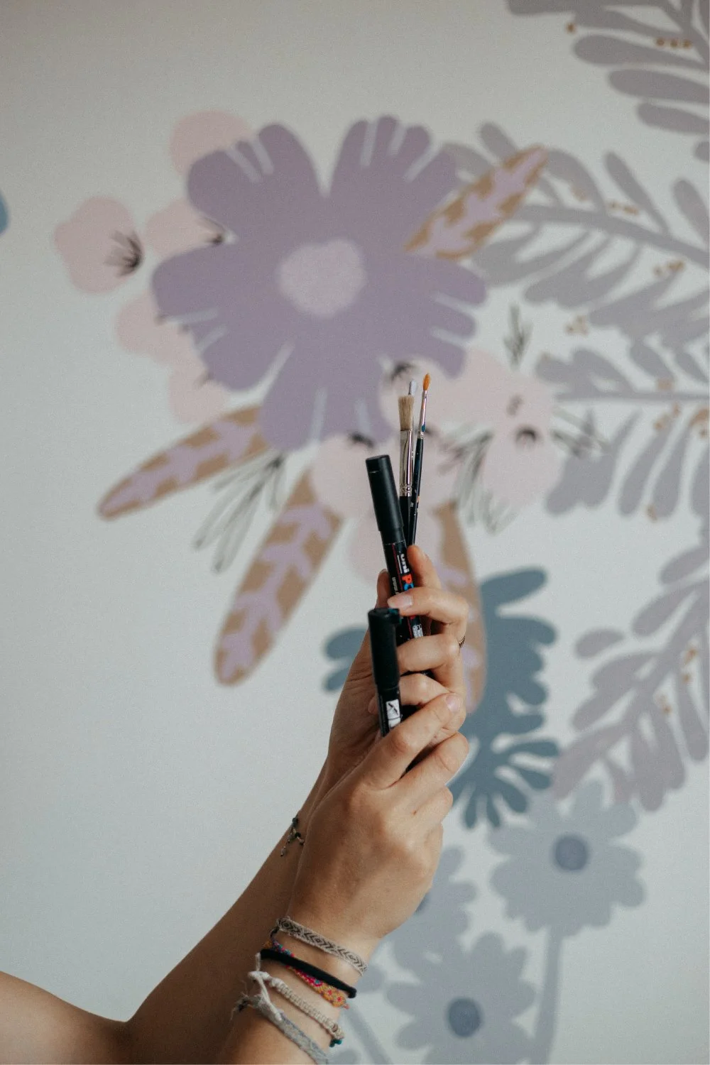 Une main tient plusieurs pinceaux devant un mur decorated avec un grand dessin floral pastel, une décoration murale.
