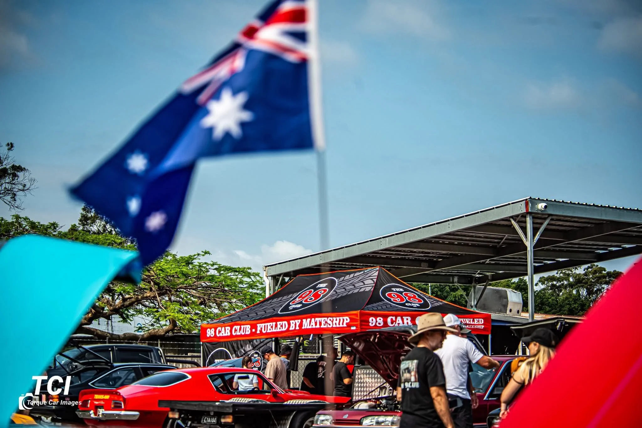 People at a car show with classic and race cars under a tent that reads '98 Car Club - Fueled by Mateship,' with Australian flags in the foreground and trees in the background.