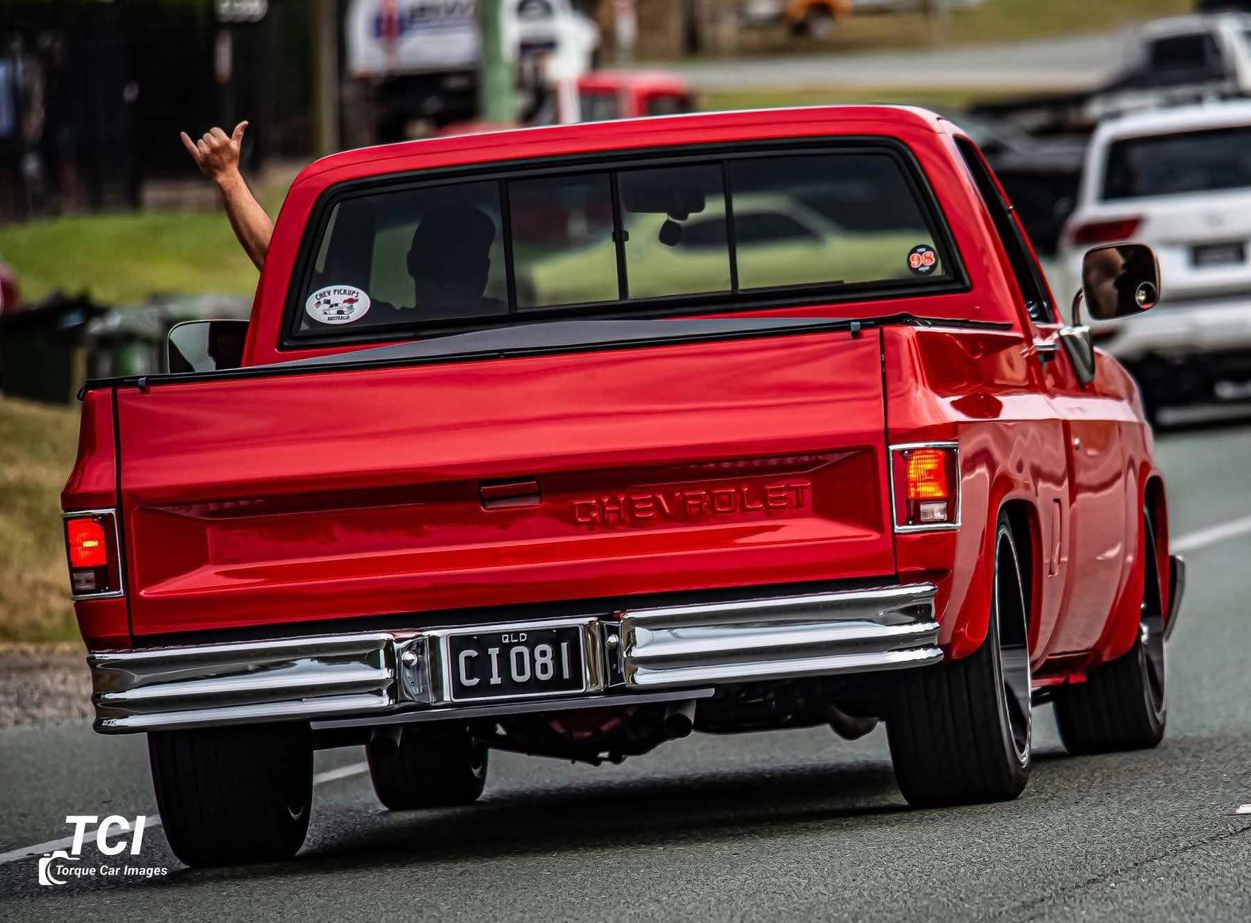 A red Chevrolet pickup truck on the road, with a person inside waving their hand out of the rear window.