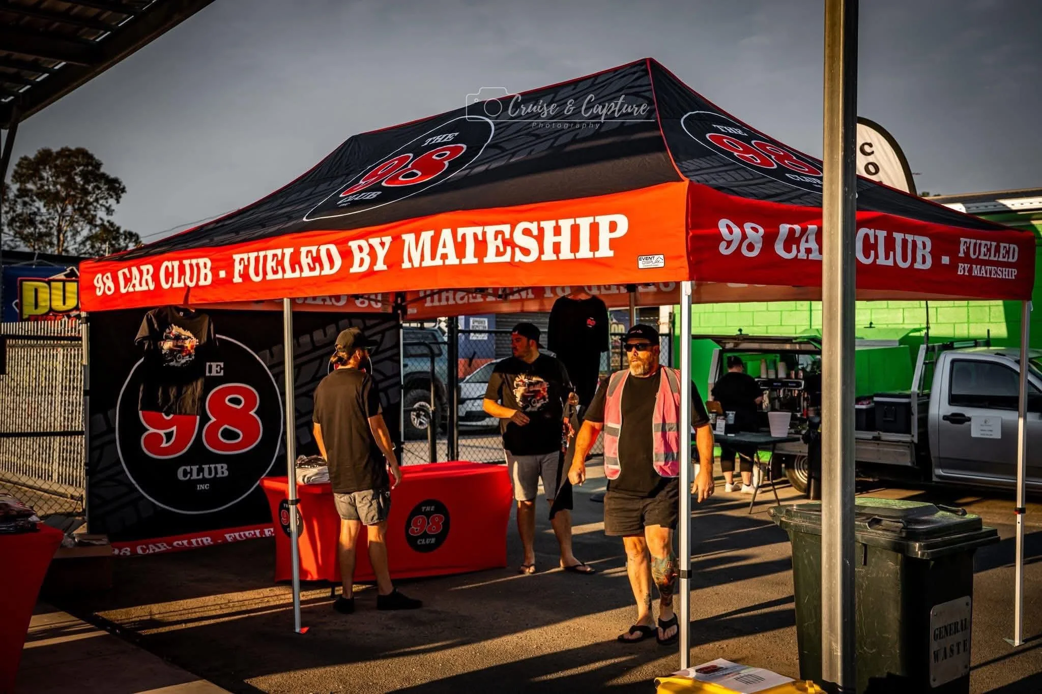 A red and black canopy tent advertising the 98 Car Club, fueled by mateship, with people gathered underneath at an outdoor event. There are vehicles and other tents in the background.