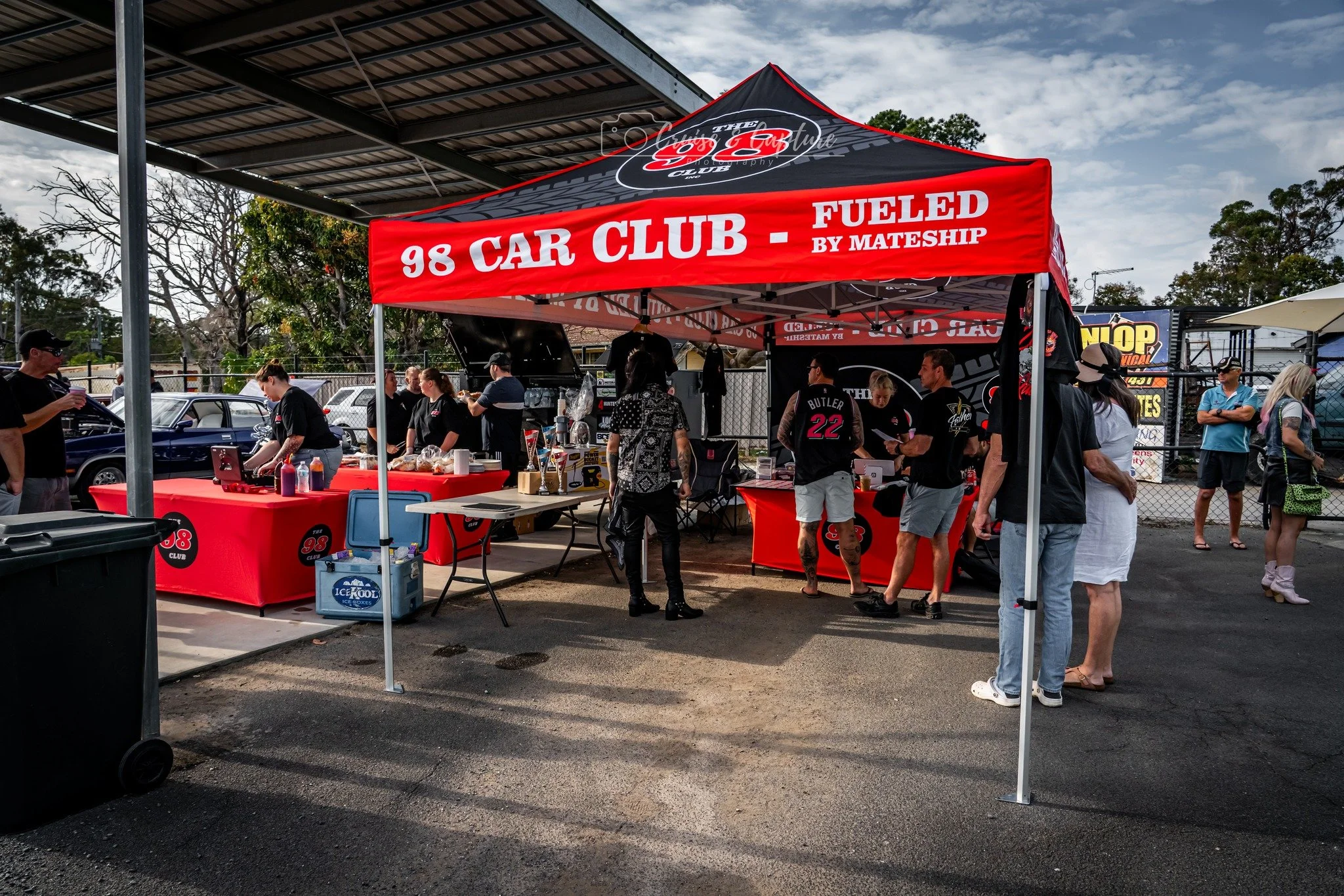 People gathered under a red and black tent with lanyards and shirts displaying '98 Car Club' at an outdoor event, with cars parked and trees in the background.