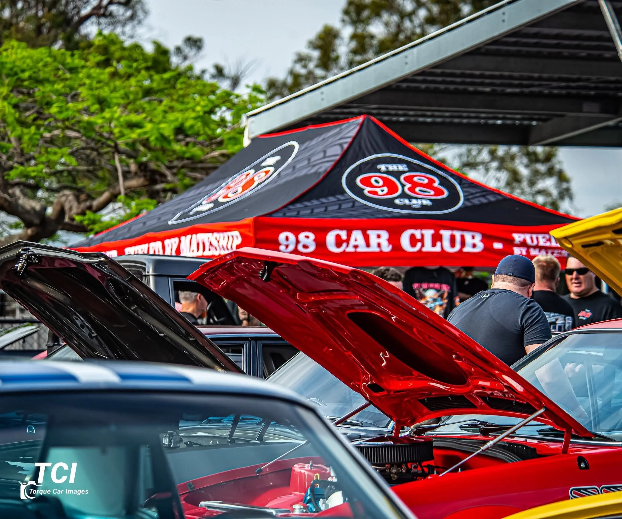 A car show with vehicles on display, including a red classic car with its hood open, showcasing the engine. There is a black tent in the background with the logo and name of the 98 Car Club. Several people are gathered around, viewing the cars and engaging in conversation.