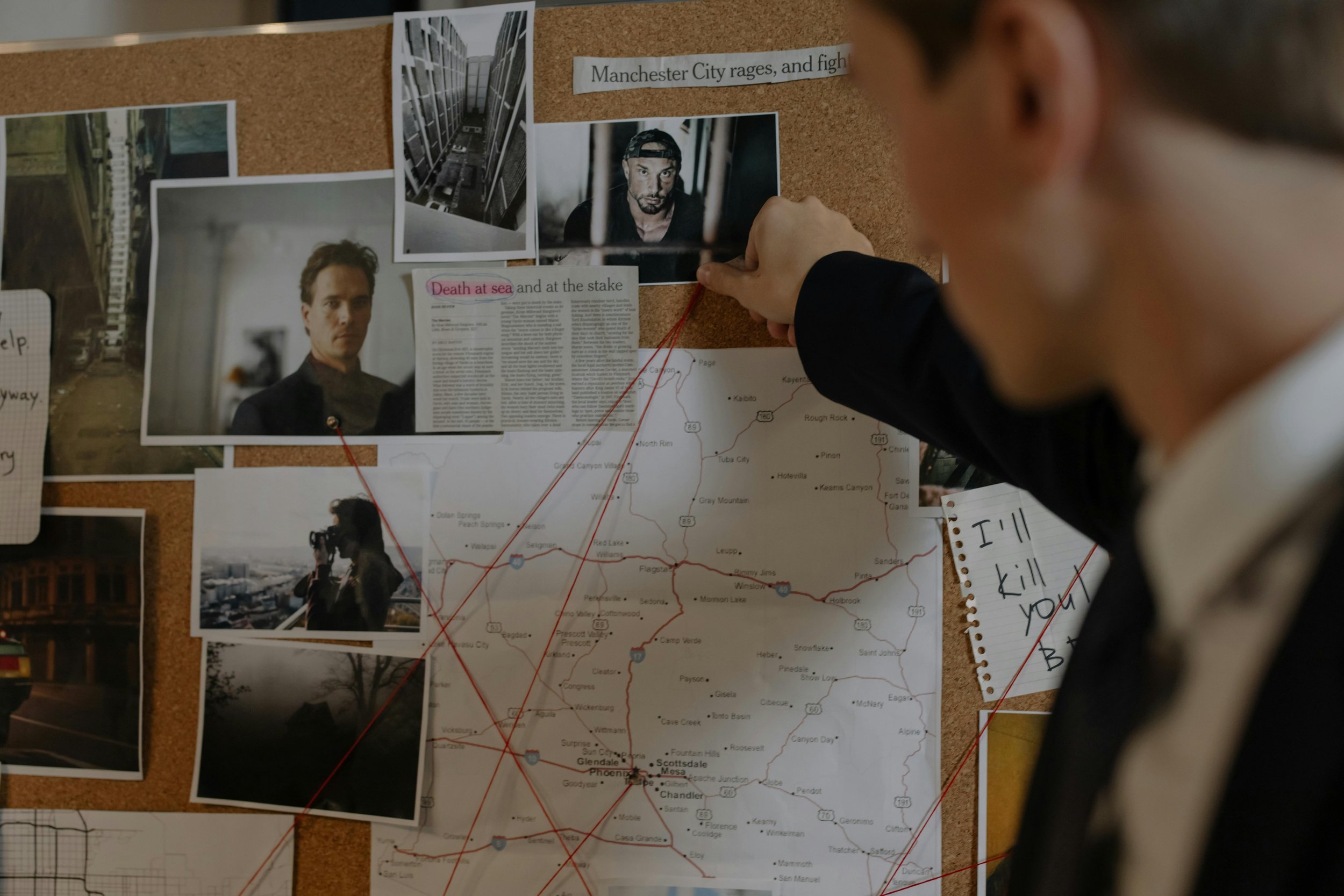 A person pointing at a bulletin board filled with photos, a map with red strings connecting various locations, newspaper clippings, and notes, suggesting an investigation or planning activity.