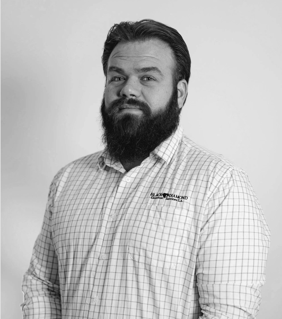 A black and white portrait of a man with a full beard and styled hair, wearing a checkered shirt with a logo that reads 'Black Diamond Electrical Services S.A.' on the left chest, standing against a plain background.