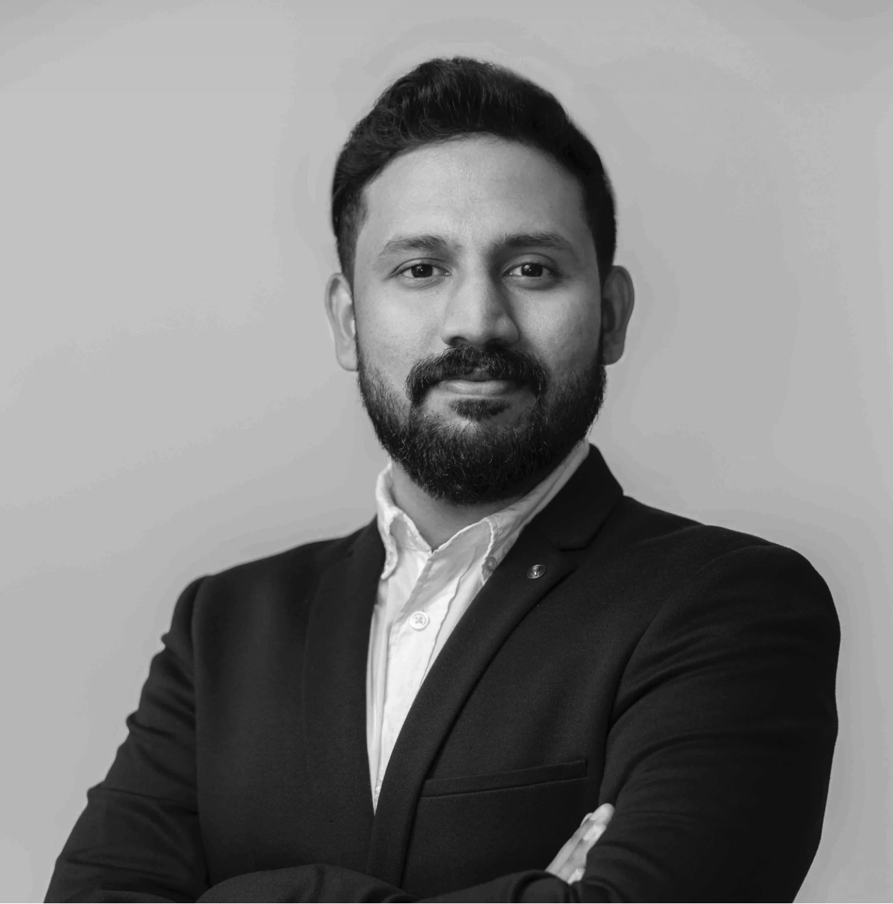 Black and white portrait of a man with a beard, mustache, and styled hair. He is wearing a dark suit jacket and a light-colored collared shirt, with arms crossed, looking confidently at the camera.
