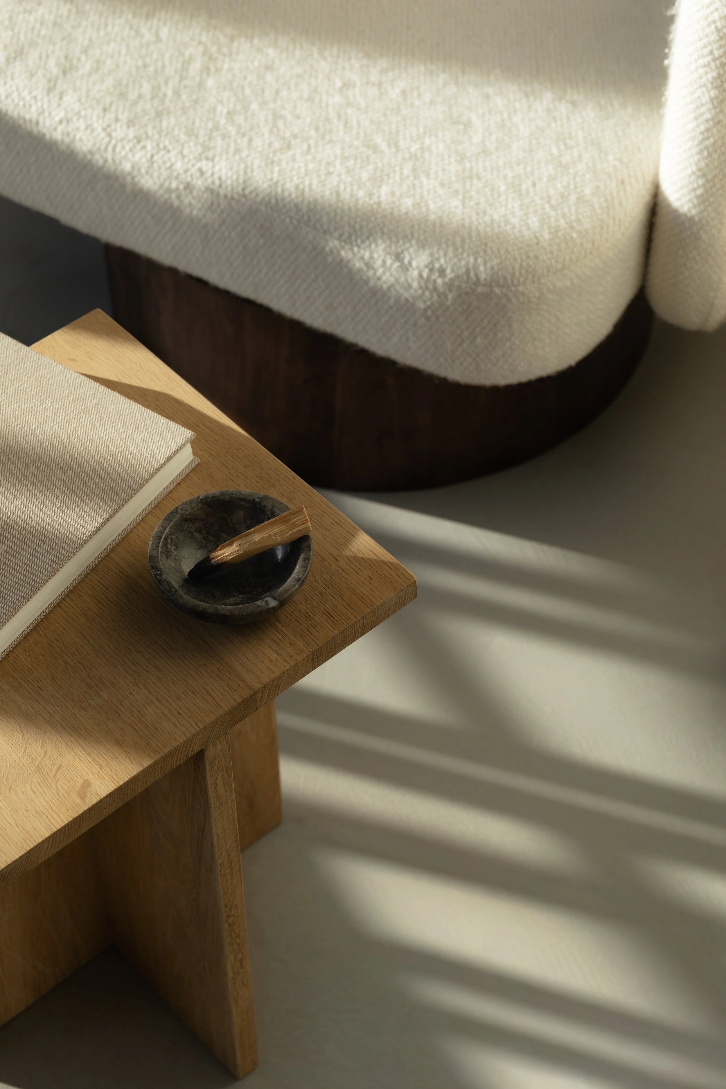 A wooden table with a black stone bowl holding a wooden stick, placed next to a stack of beige cloth napkins, with a beige upholstered chair in a sunlit room.