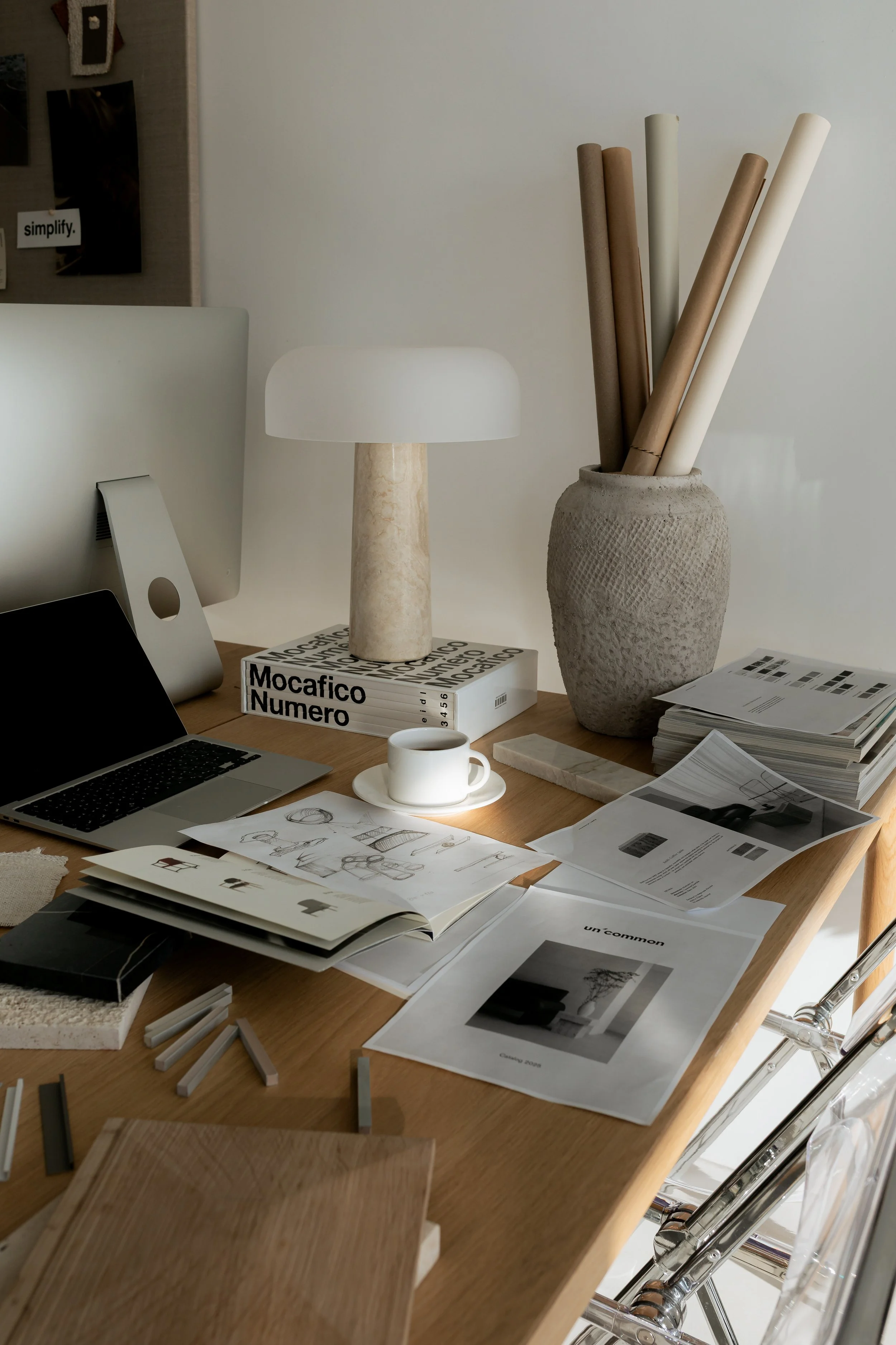 A cluttered wooden desk with a computer monitor, laptop, books, magazines, sketches, a coffee cup on a saucer, a table lamp, and a large textured vase holding rolled papers, in a modern workspace with white walls.