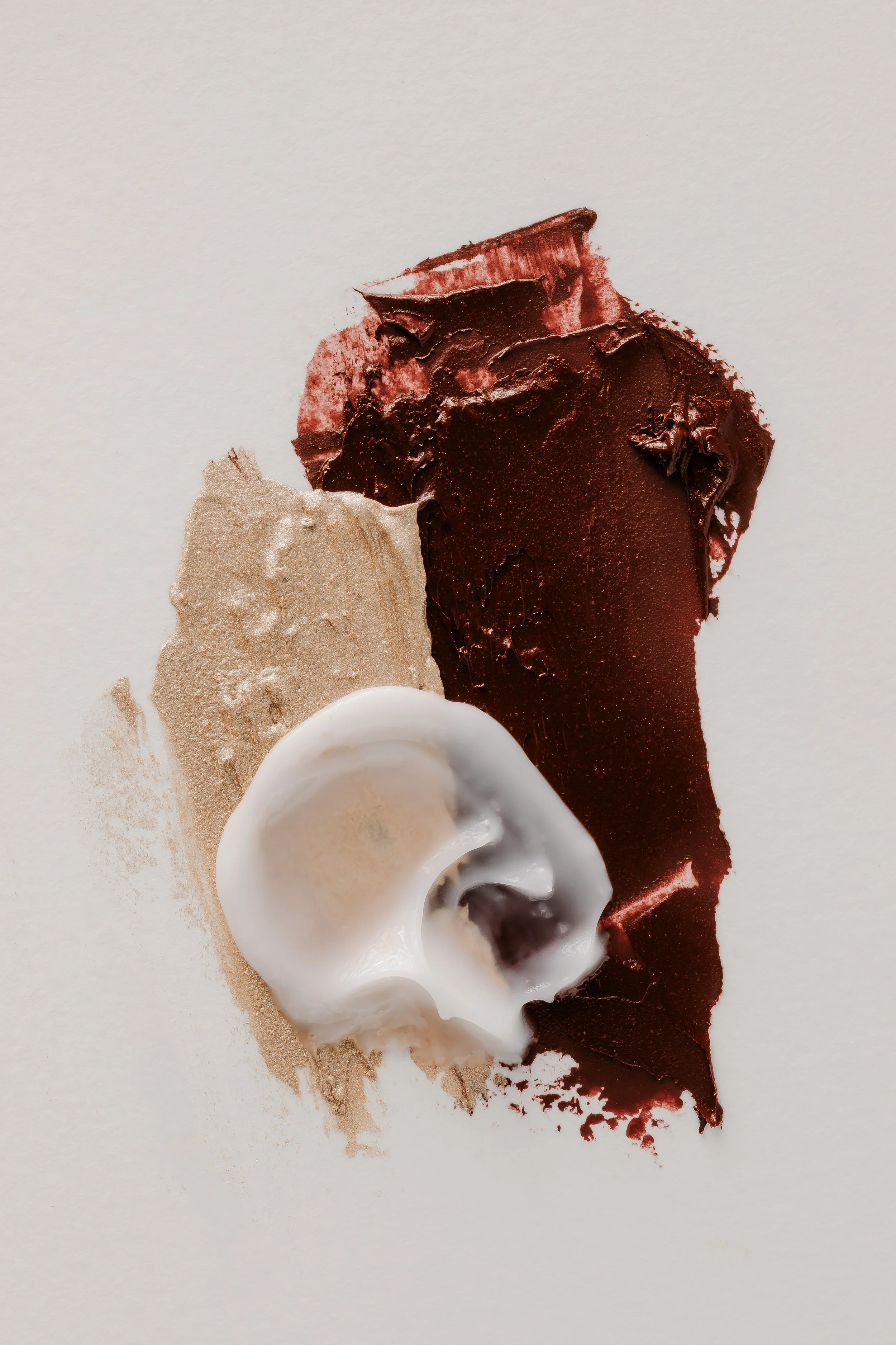A white background with swatches of beige, dark brown, and white cream, possibly cosmetics or paint.