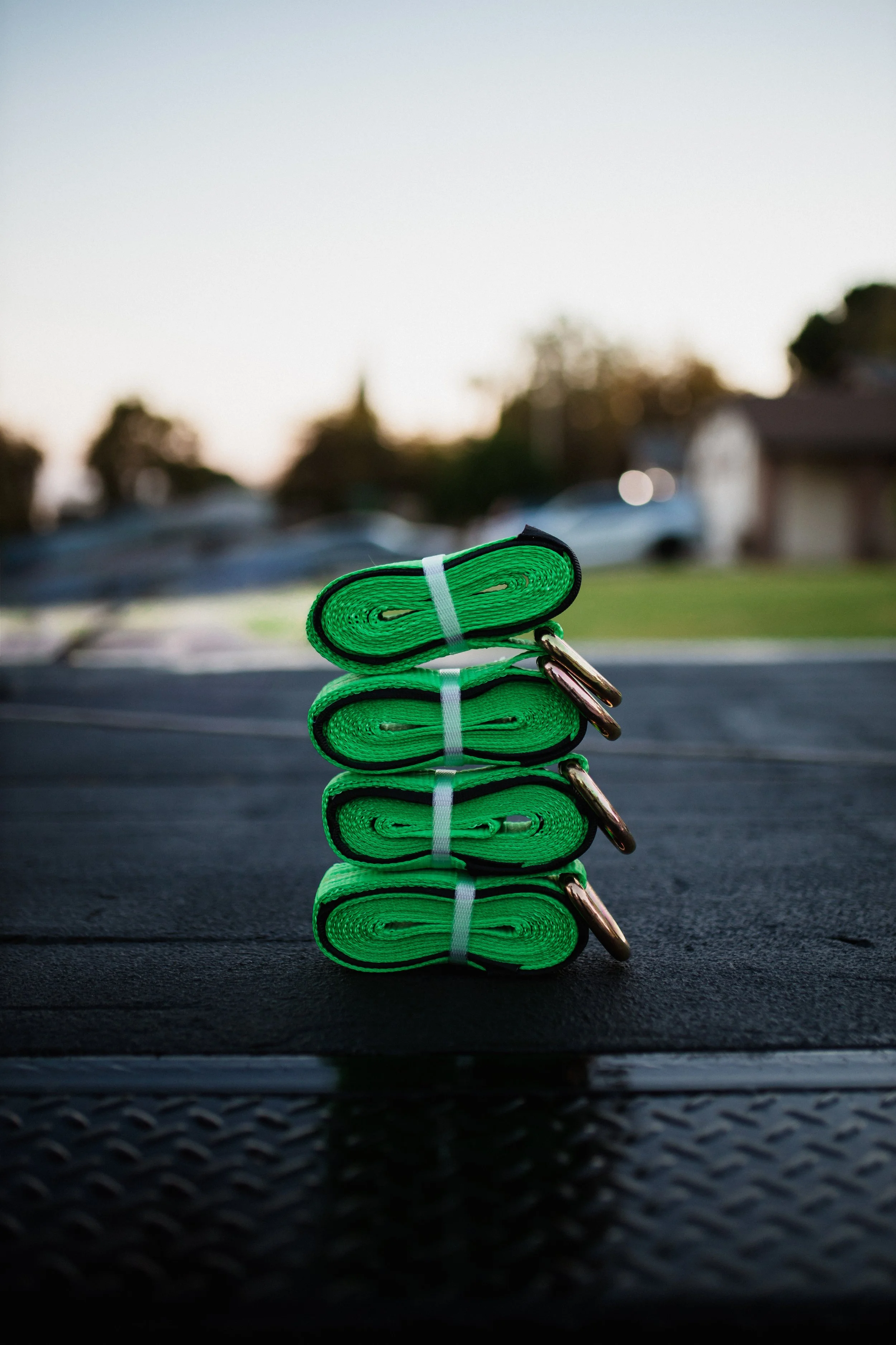 Stacked green resistance bands with metal handles on a black surface outdoors, with a blurred suburban background during sunset.