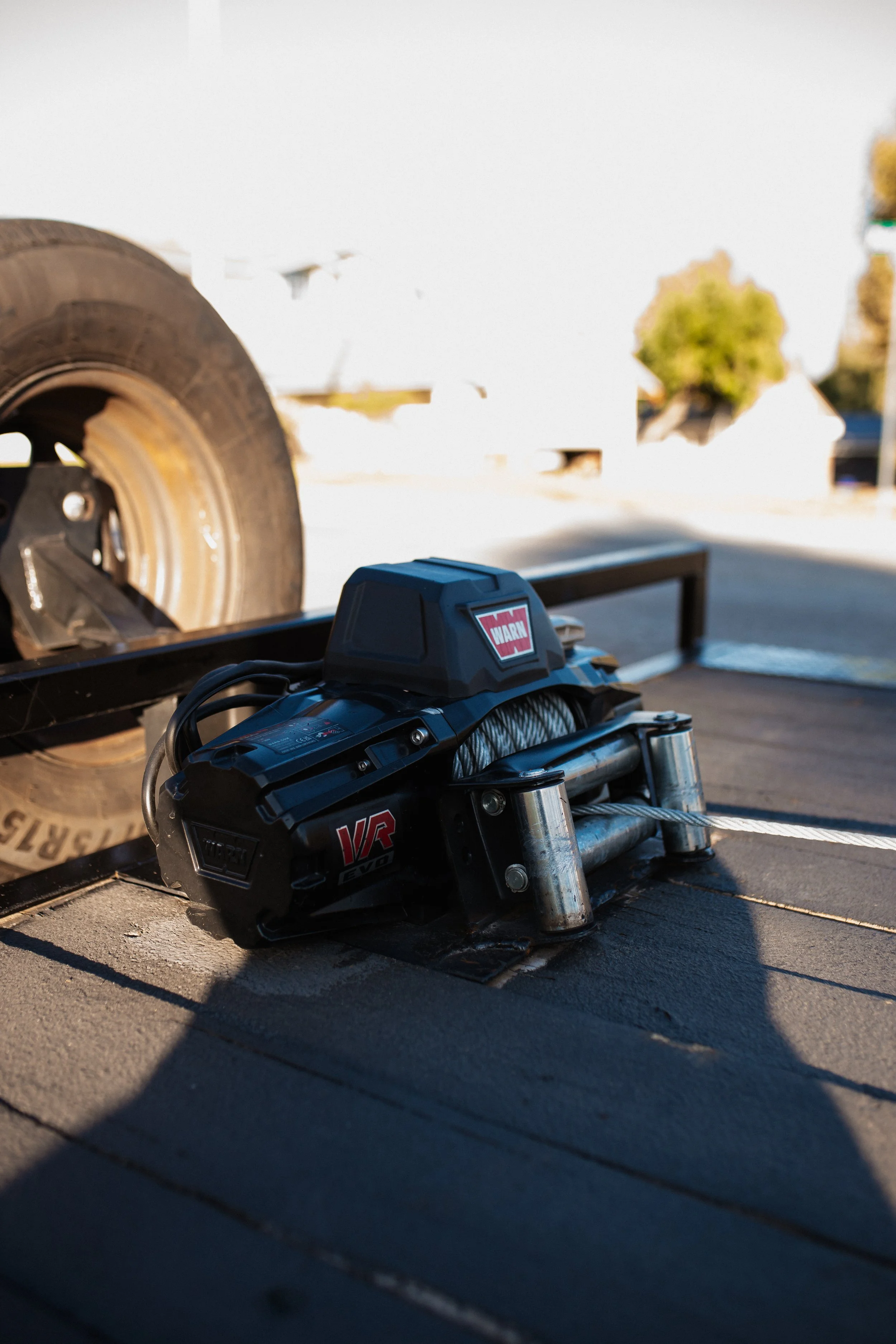 Close-up of a black electric winch with cables, mounted on a flat surface, with a vehicle tire partially visible in the background.