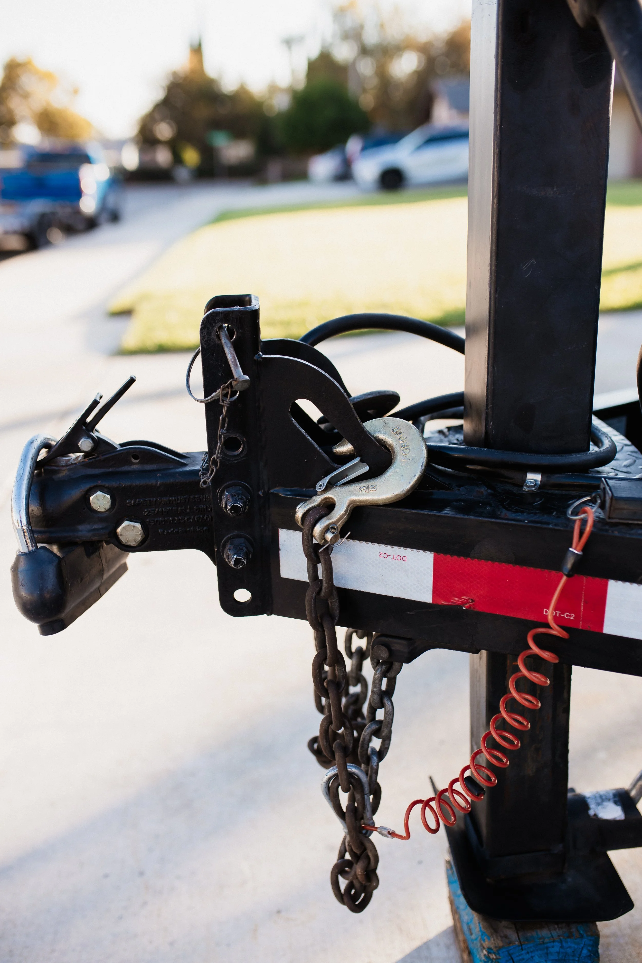 Close-up of a trailer hitch secured with a chain and lock, with a red coiled air brake line attached, on a black trailer frame parked outdoors.