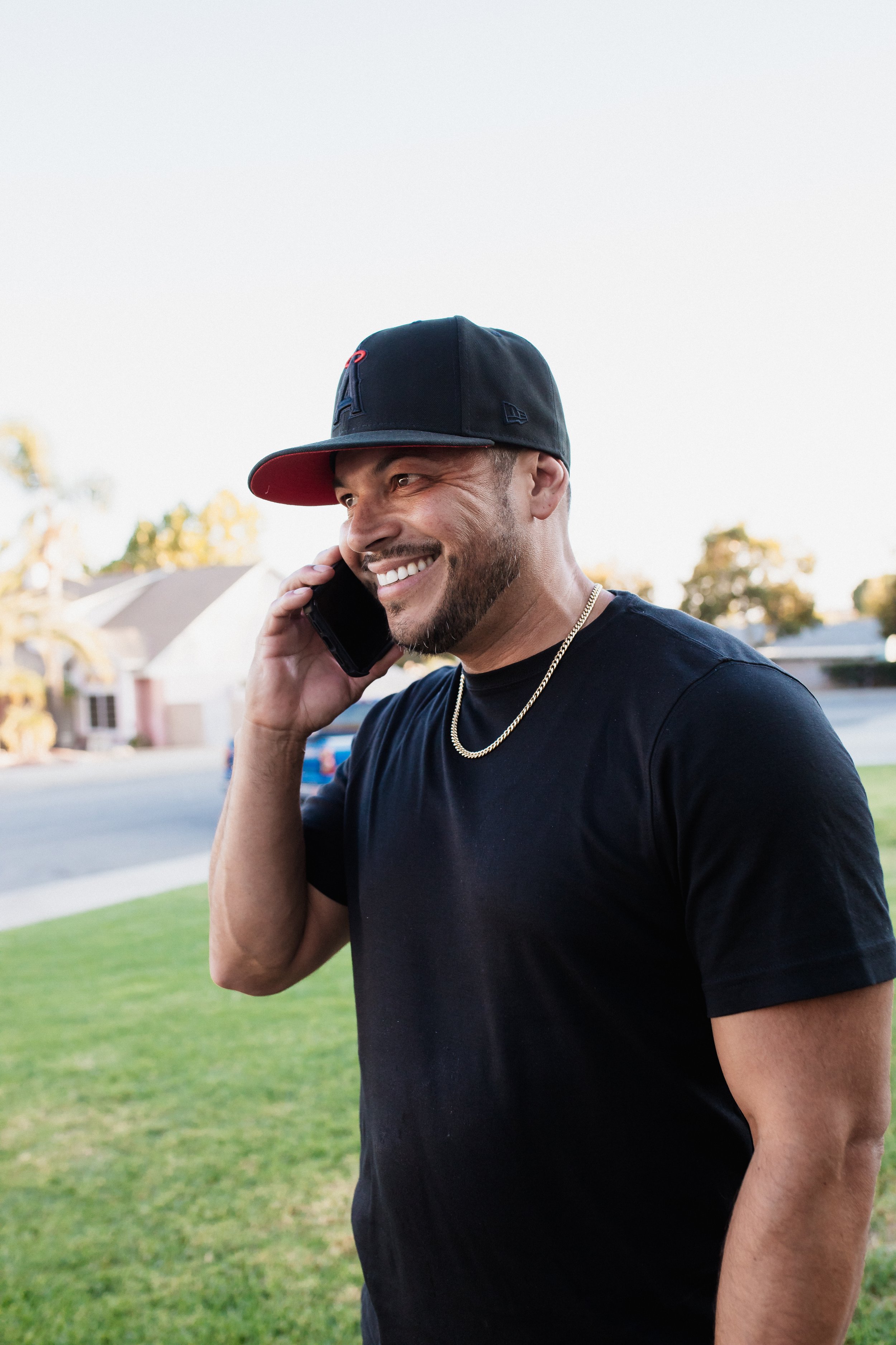 A man wearing a black baseball cap with a red brim, a black t-shirt, and a gold chain is smiling and talking on a mobile phone outdoors in a suburban neighborhood with houses and trees in the background.