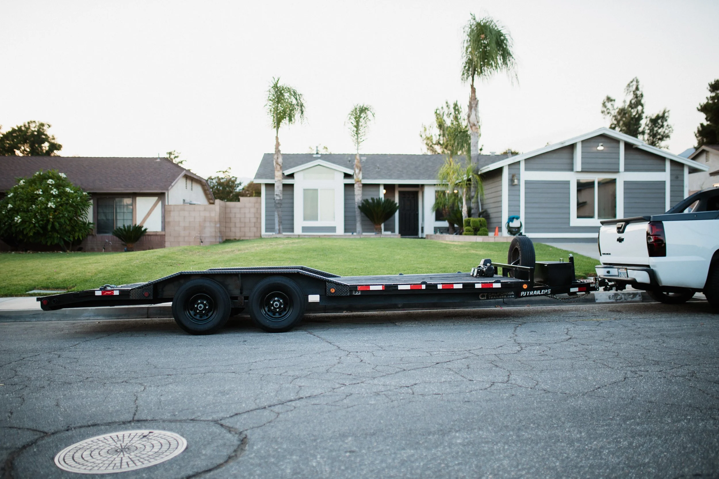 A white pickup truck with a trailer attached, parked on a residential street in front of a house with gray siding and palm trees.