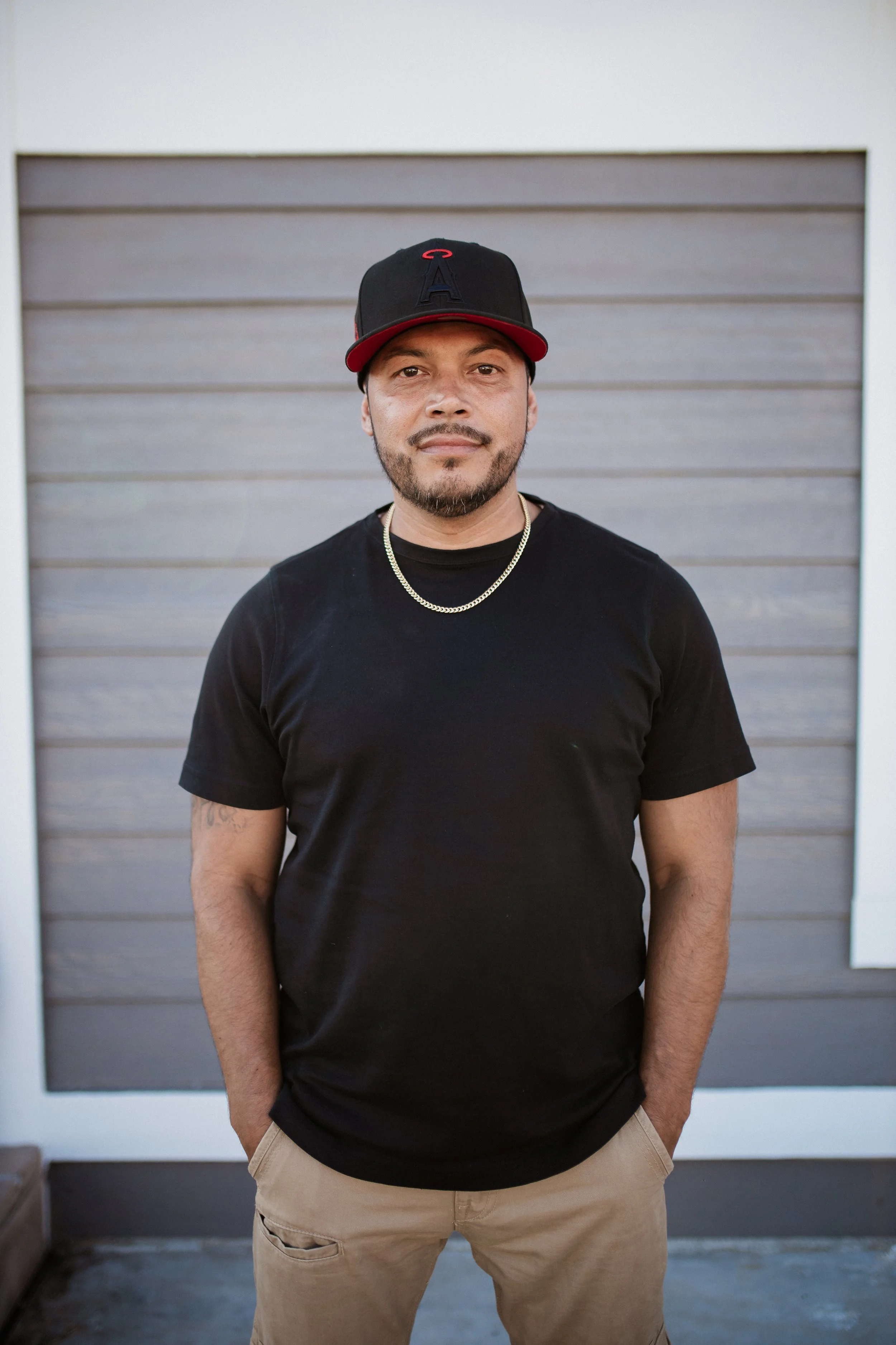 A man with a beard and mustache wearing a black baseball cap, black t-shirt, and gold chain necklace standing outdoors in front of a brown garage door.