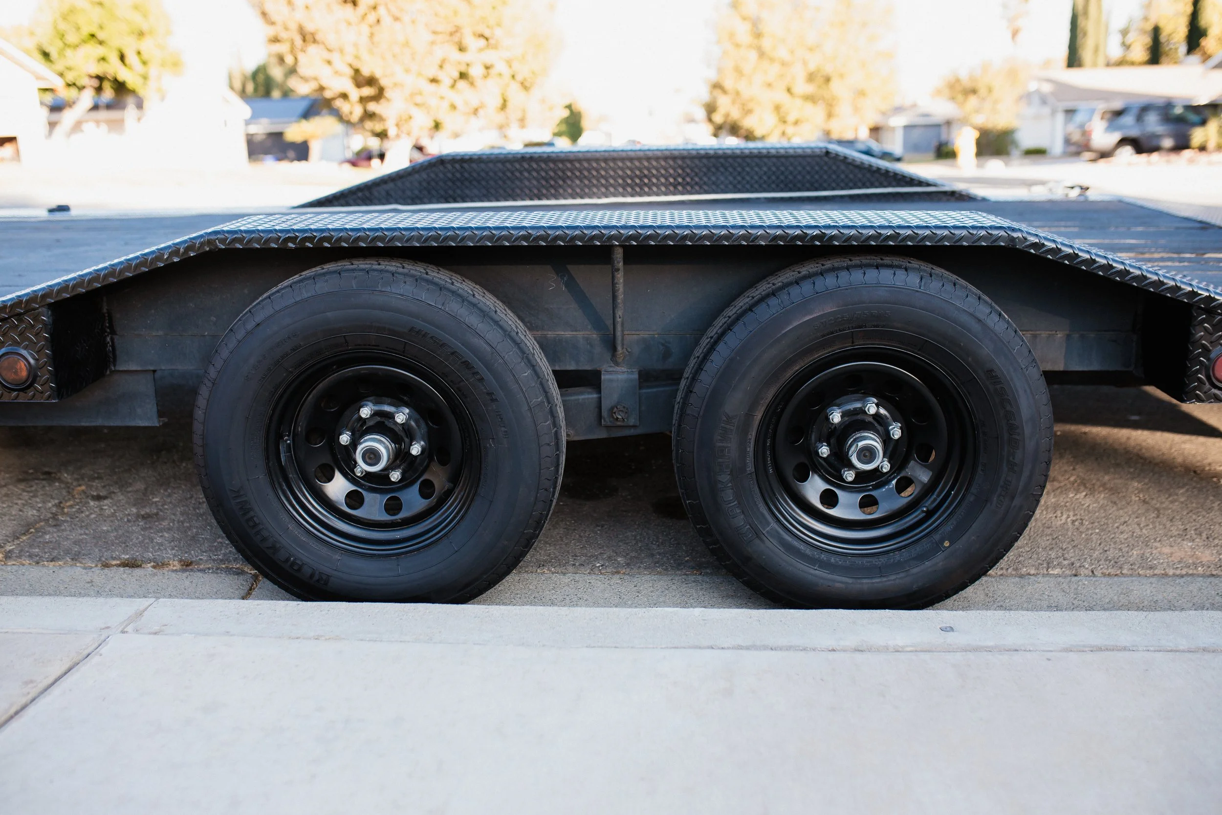 A dual-axle black trailer parked on a driveway, with a flat metal surface and black tires with silver bolts.