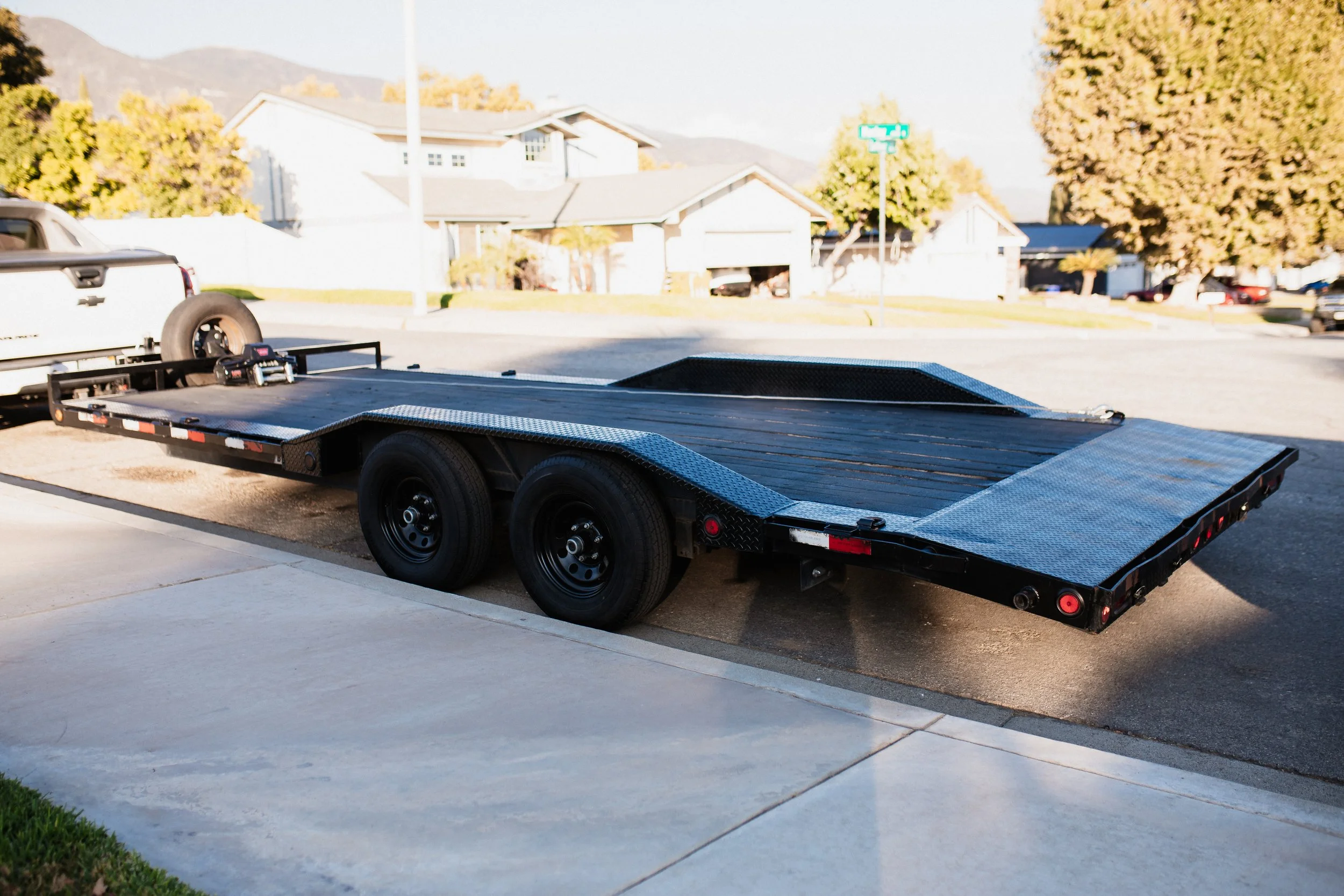 A black car trailer parked on a driveway in a residential neighborhood, with a white truck attached at the front of the trailer.