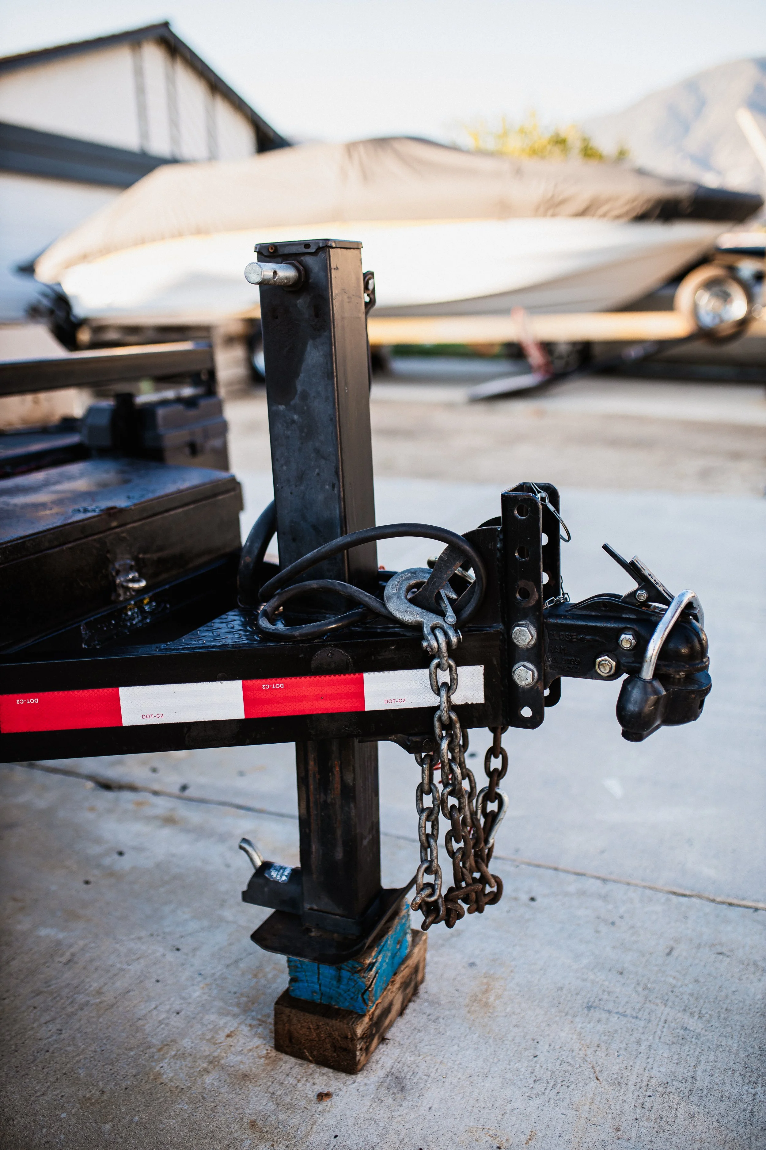 Close-up of a trailer hitch with chains, parked on a concrete surface, with a boat covered with a tarp in the background.