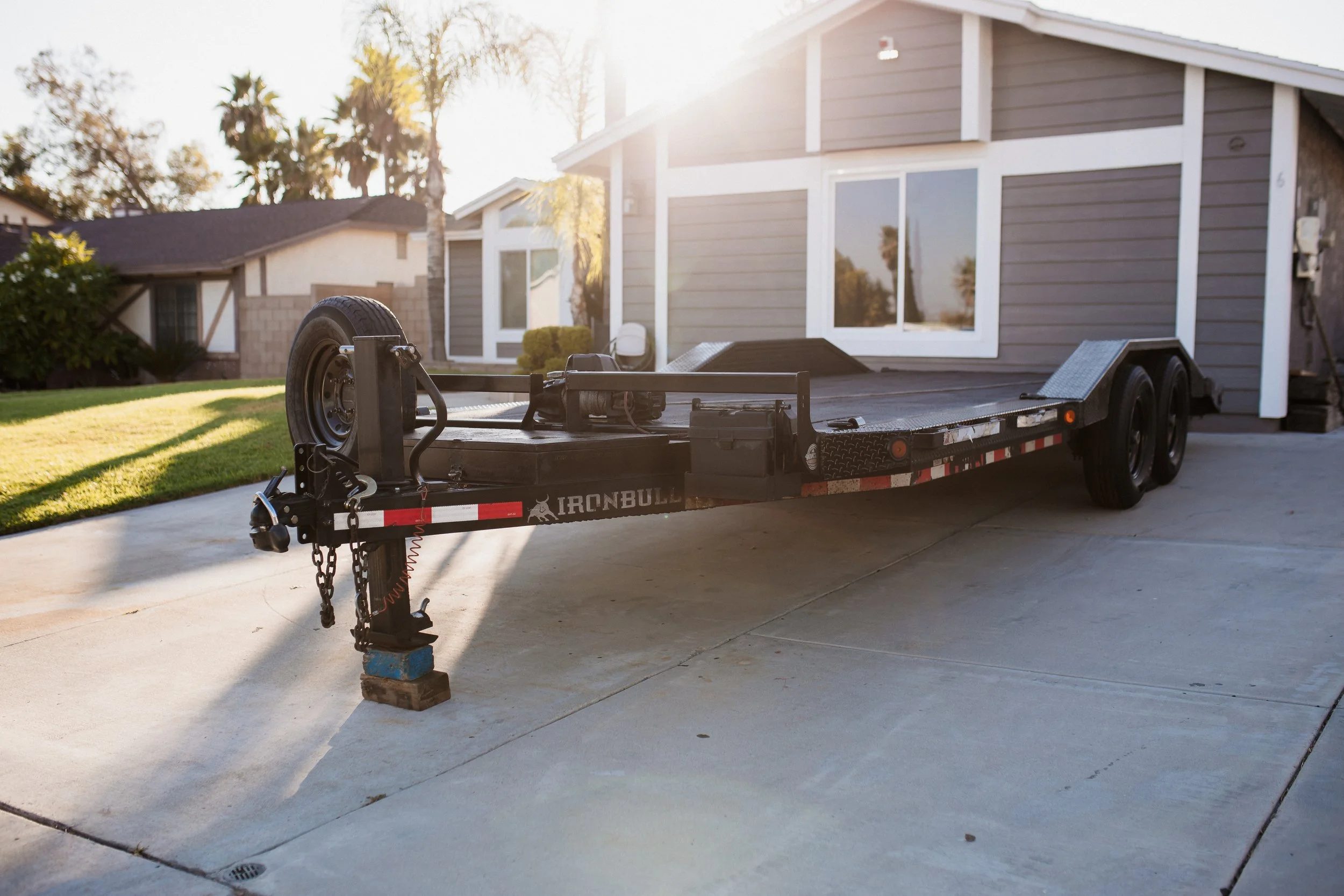 A trailer parked on a driveway in front of a house with grey siding, large windows, and palm trees in the background, illuminated by sunlight.