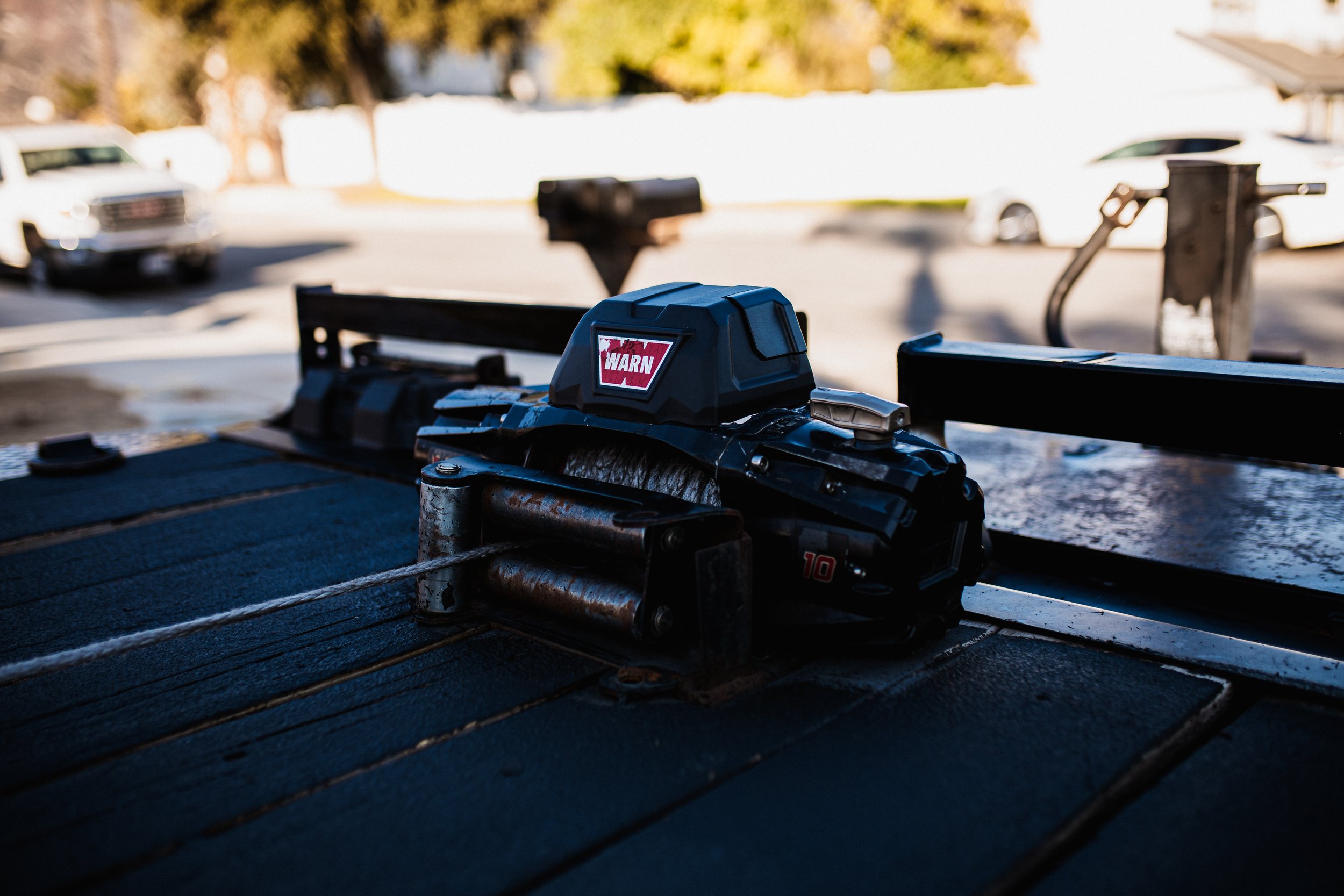Close-up of a trailer winch on a black wooden surface, with a warning label on the top, outdoors with cars and trees in the background.