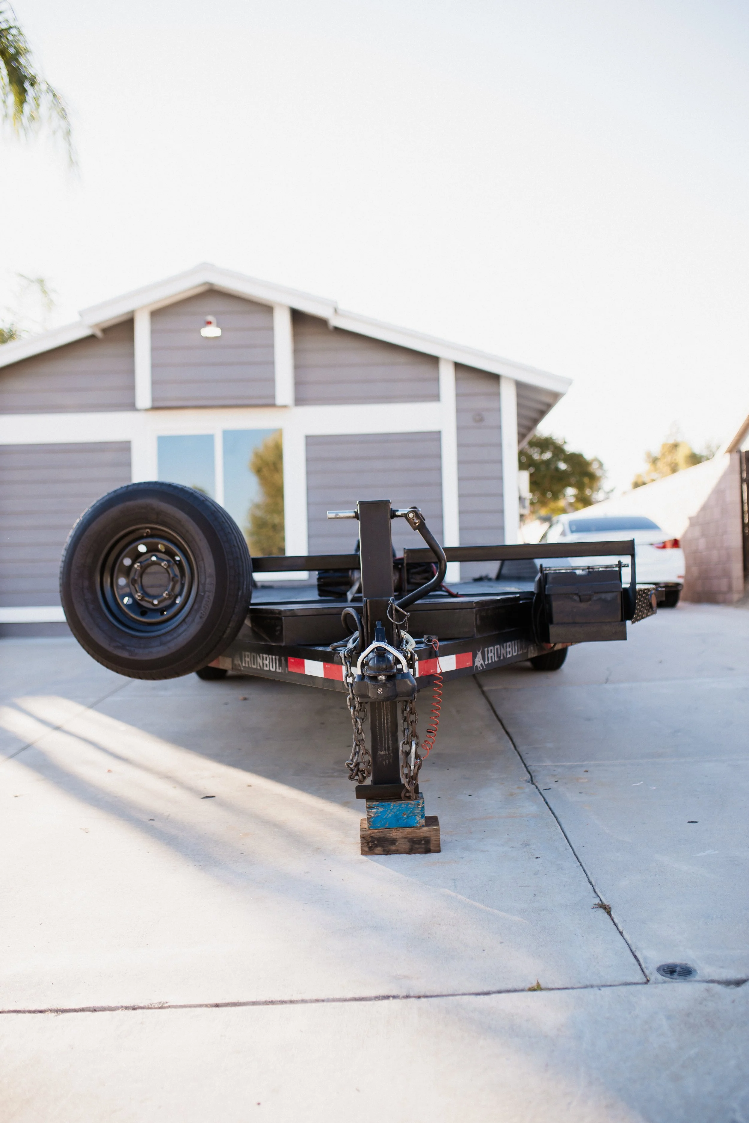 A flatbed trailer with a mounted spare tire, parked on a driveway in front of a house with gray siding and large windows.