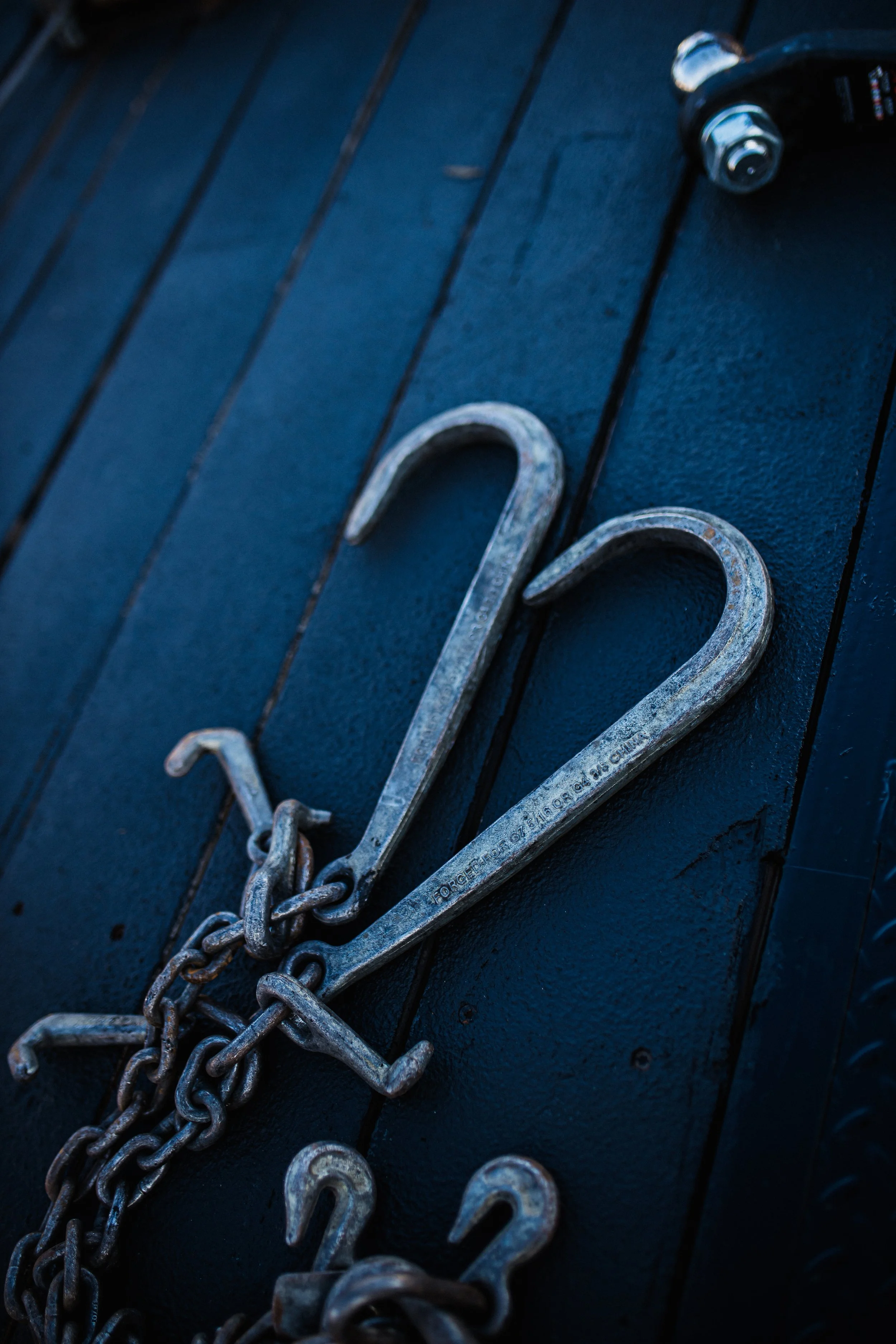 Close-up of a metal hook shaped like the number 12, chained to a dark blue wooden surface, with a small padlock and chains nearby.