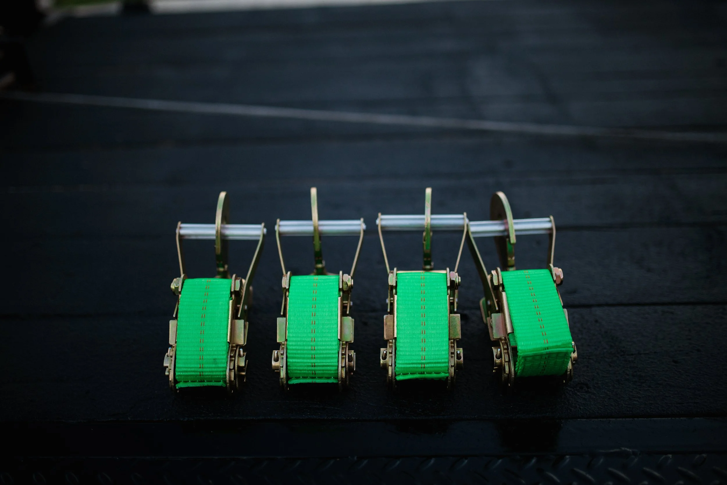 Four green racing harnesses connected to a metal bar, used to secure a driver in a race car, set against a dark background.