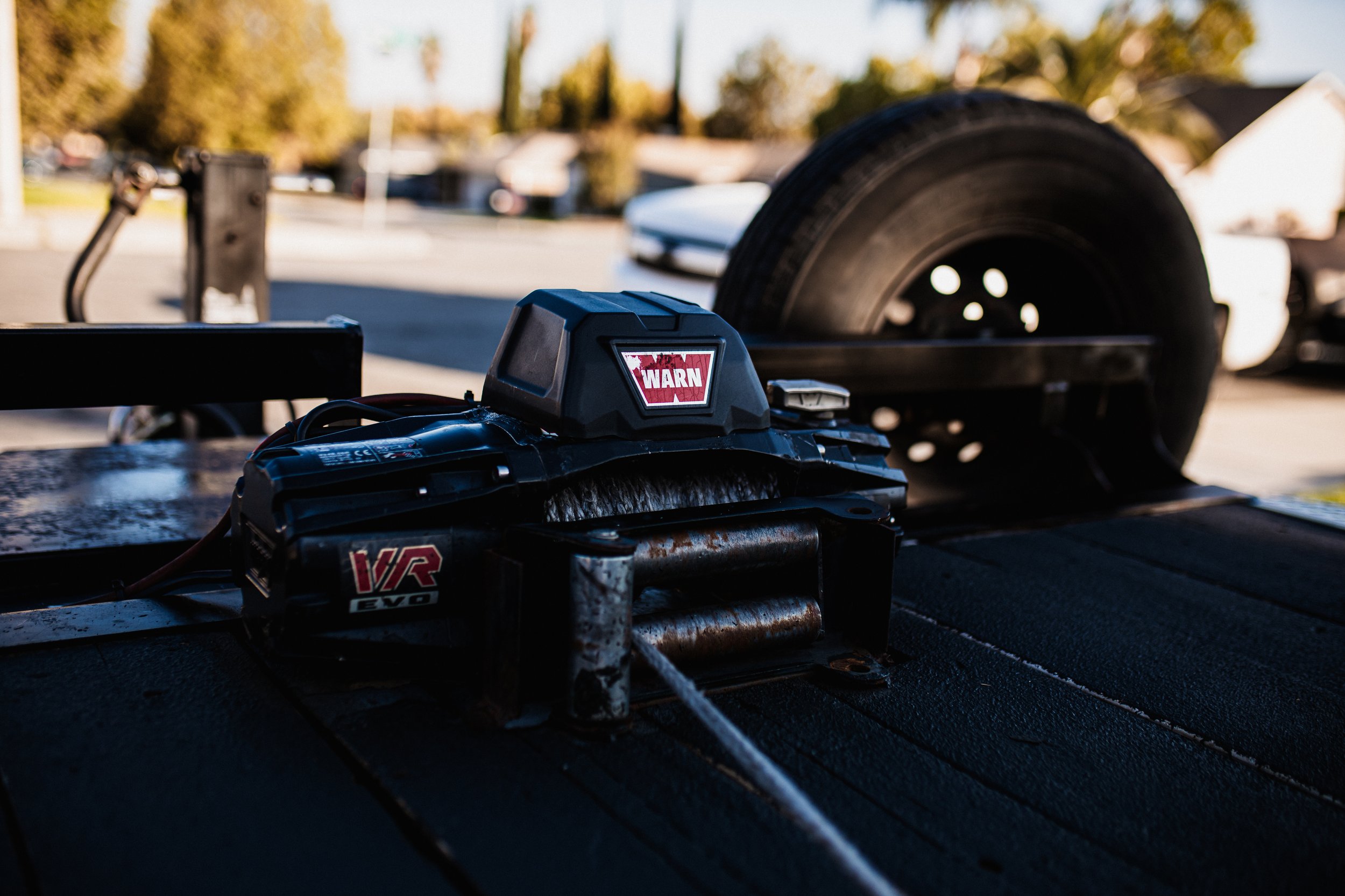 Close-up of a winch with a warning sticker, mounted on a flat surface, with a trailer wheel in the background, outdoors in daylight.