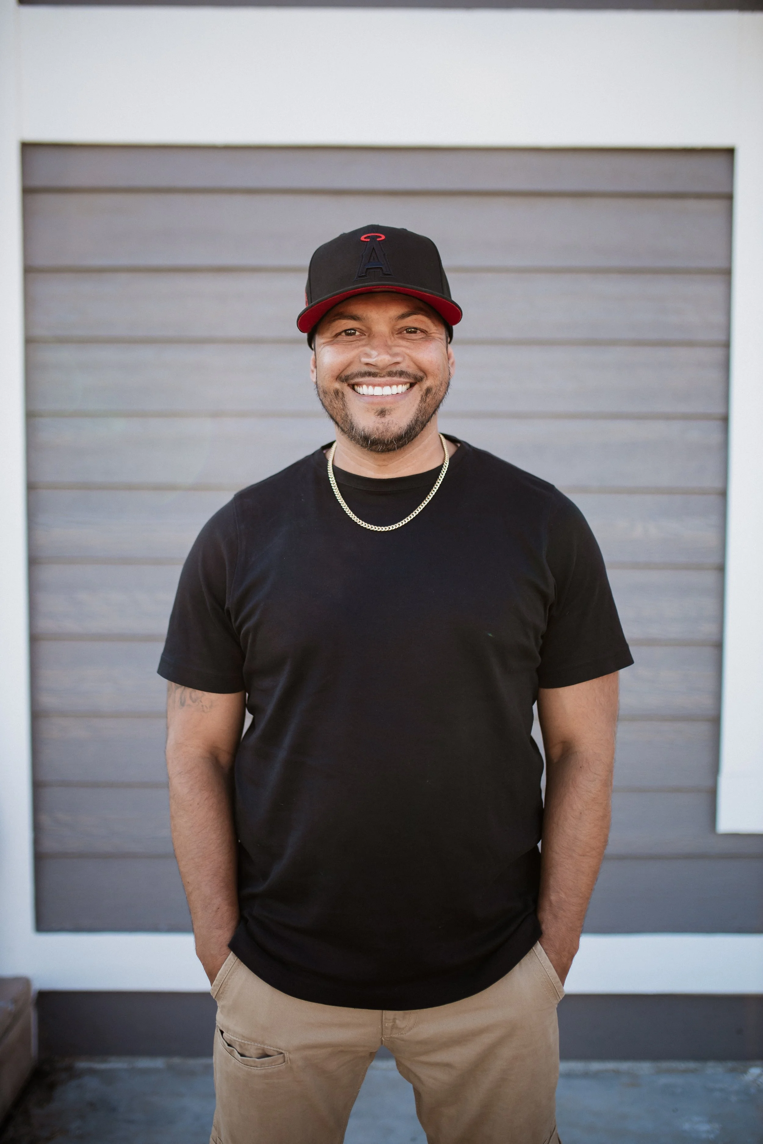 A smiling man outdoors standing in front of a gray and white building, wearing a black t-shirt, beige pants, a black and red baseball cap, a gold chain, with hands in pockets.