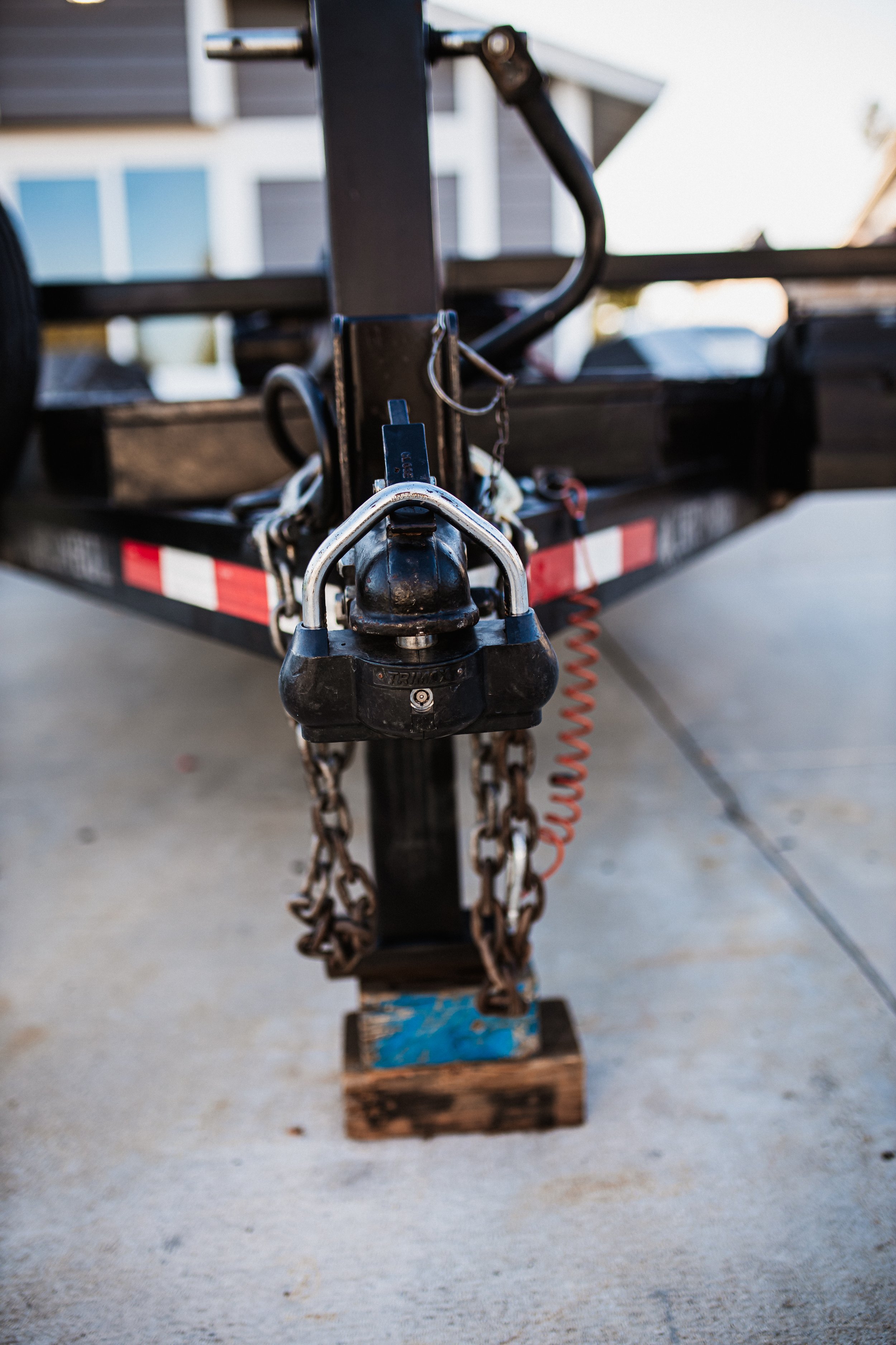 Close-up of a trailer hitch lock, chains, and safety chains attached to a flatbed trailer, with a house in the background.