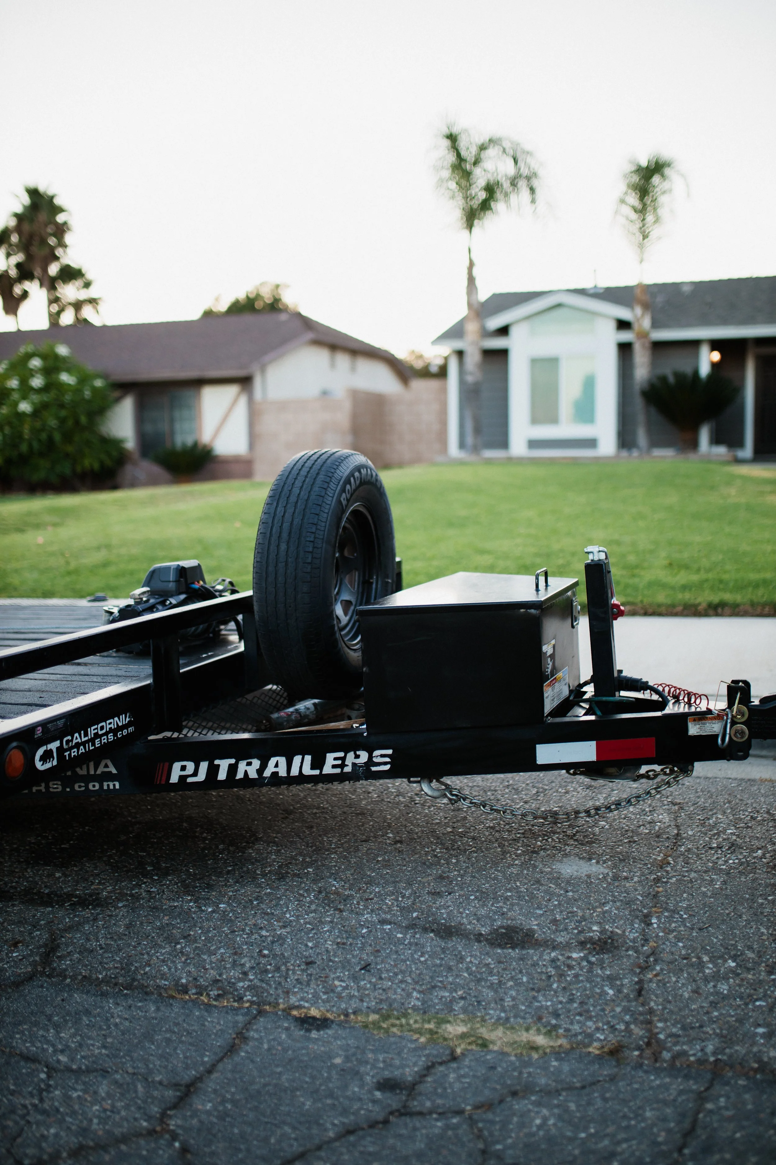 A black trailer with a spare tire, parked on a driveway in front of a suburban house with a lawn and palm trees.