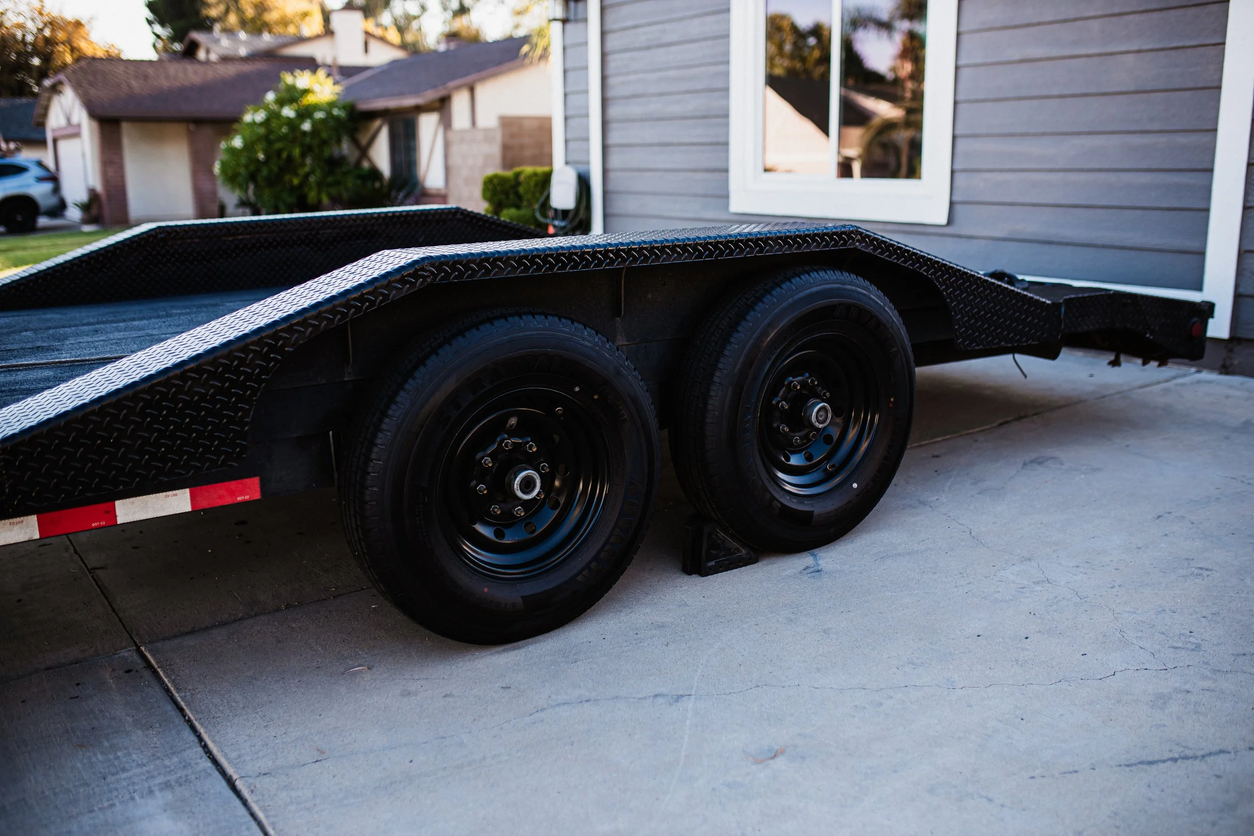 A black flatbed trailer parked on a concrete driveway in front of a house with gray siding and a white window. The trailer has two wheels on each side and is propped up with a wheel chock.