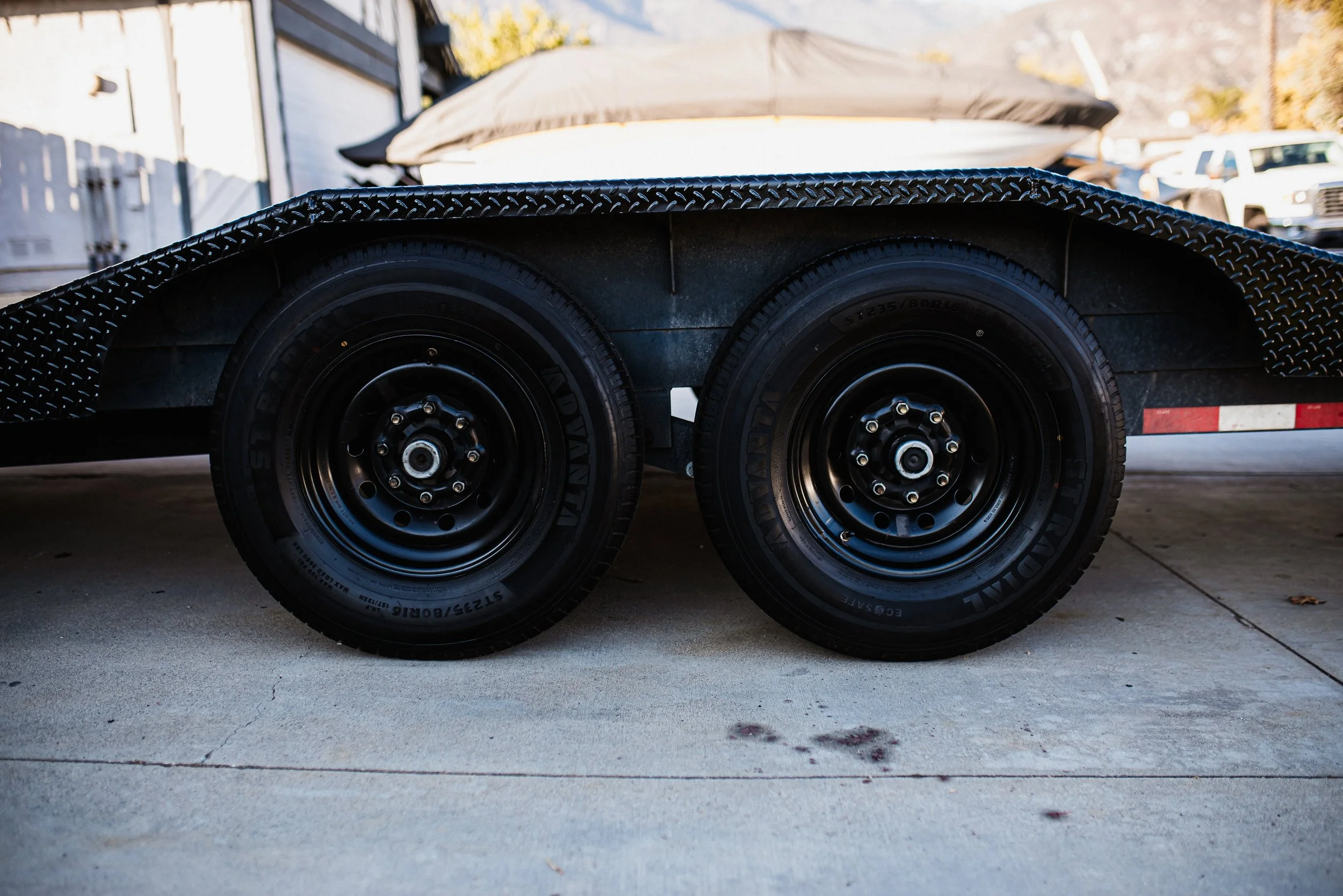 Close-up of a dual-axle boat trailer with black tires, parked on concrete, with a boat covered in a tarp and some cars in the background.