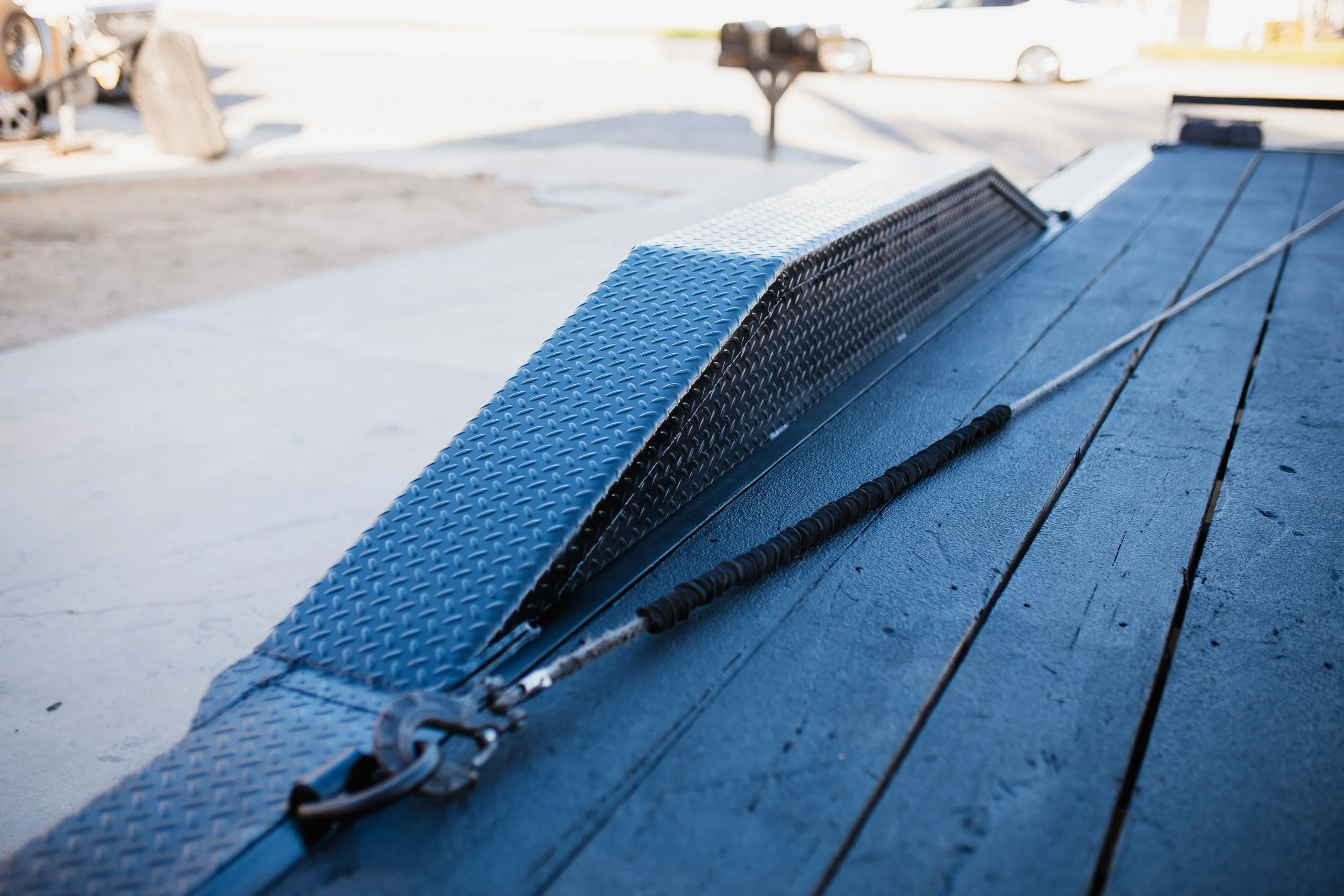 Close-up of a flatbed trailer's deck with a metal ramp and a tensioning bar, outside parking lot with vehicles in the background.